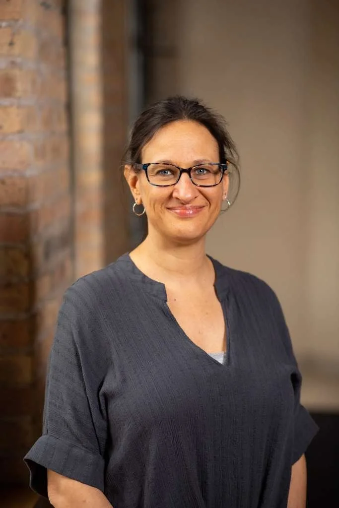 Rehabilitation Therapist, Doris Martinkus, stands against a brick background smiling and wearing a navy blue top.