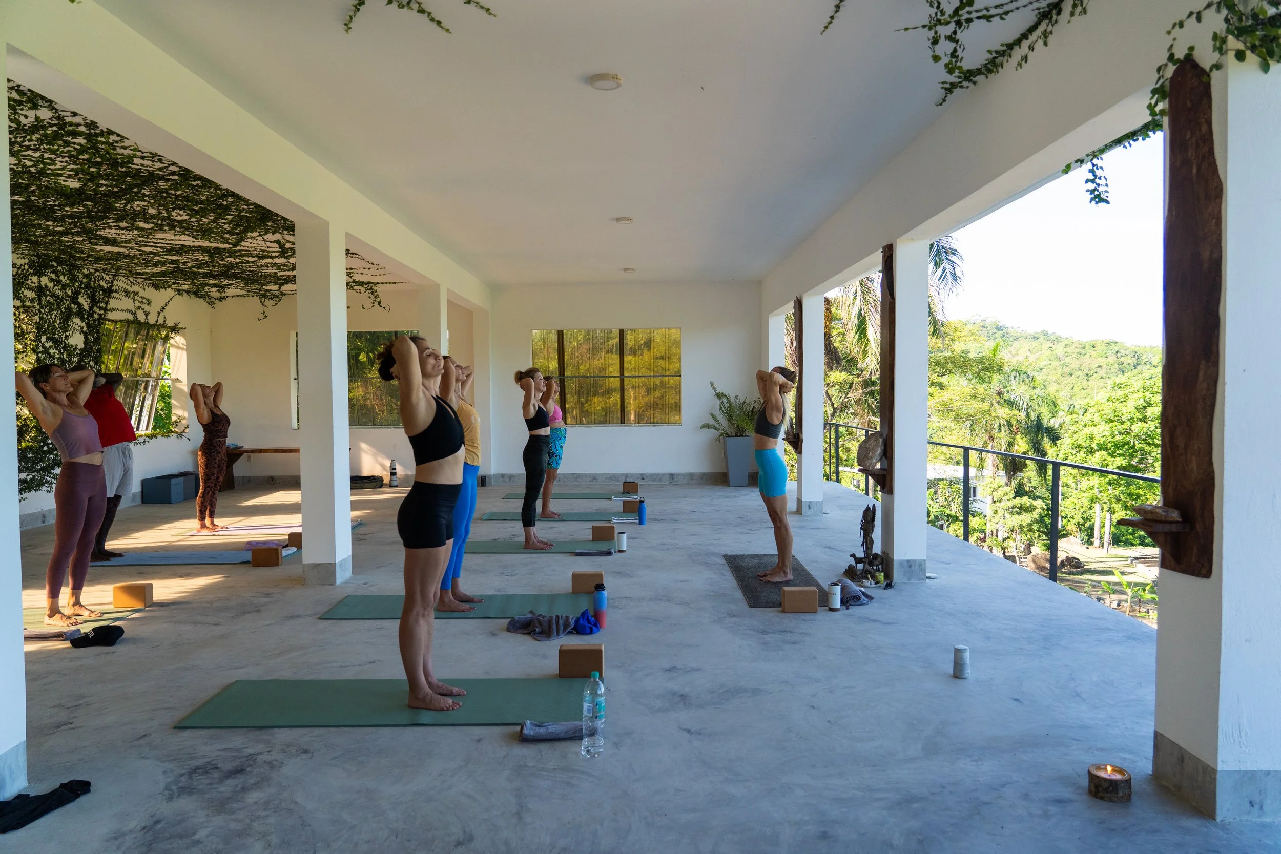 A group of women participating in a yoga or meditation class on a spacious balcony with a view of lush green trees, some with their hands behind their heads, standing on yoga mats with water bottles and props nearby.