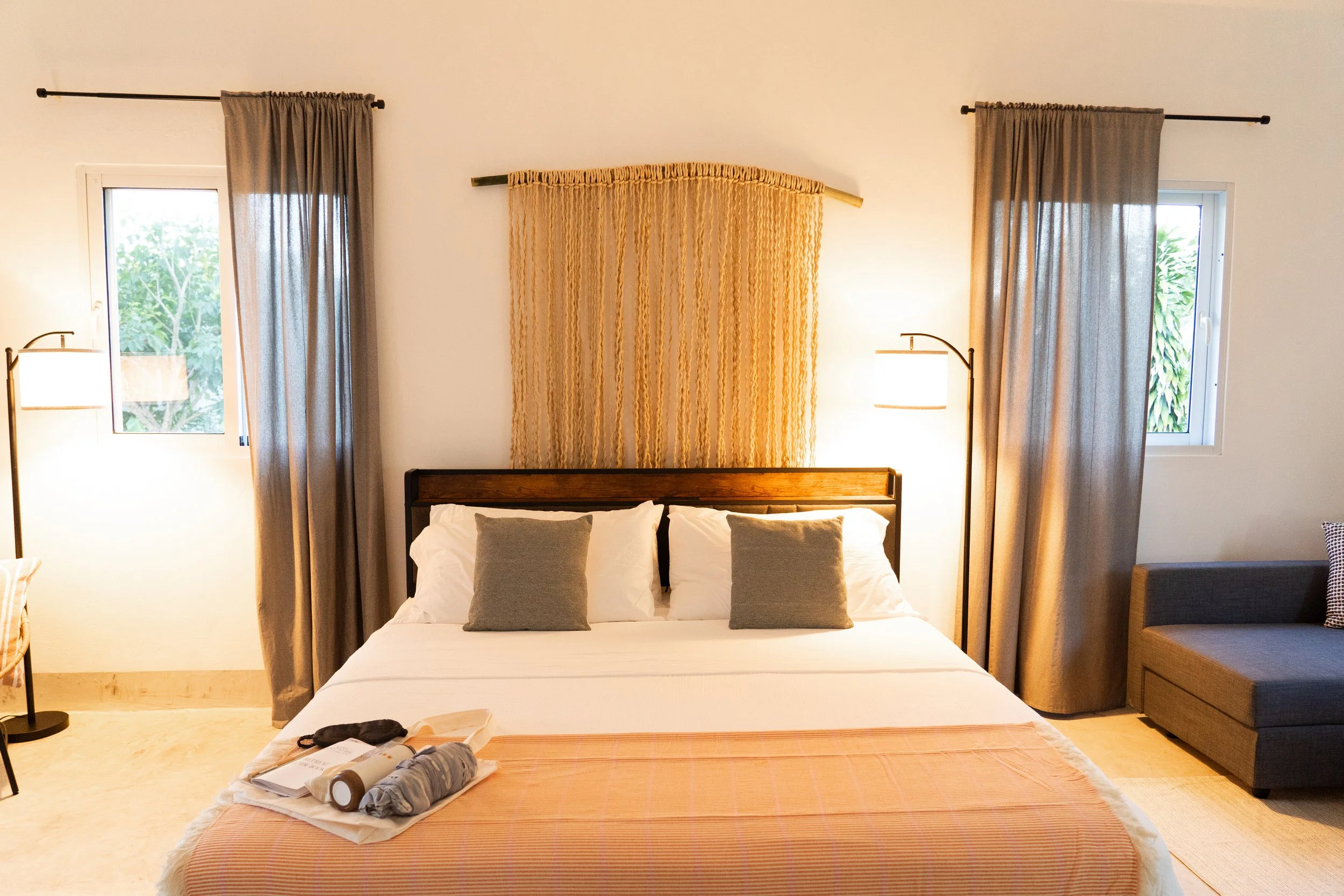 Hotel bedroom with a bed, two windows with gray curtains, a dark wood headboard, two lamps, and a gray sofa on the right side.