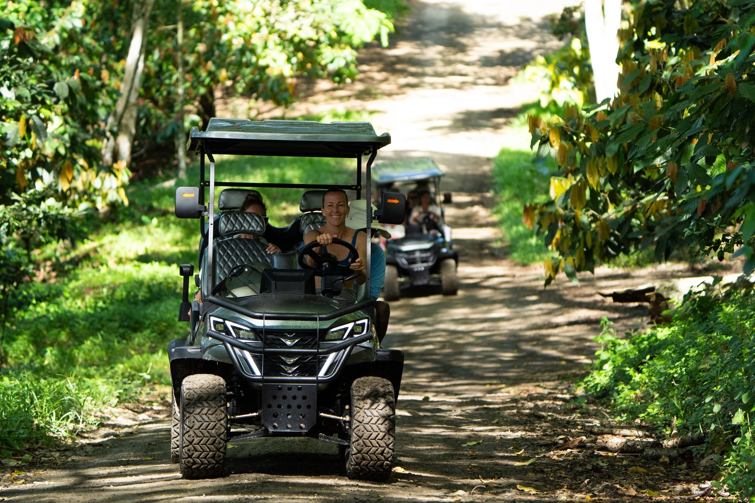 People riding in black golf carts on a dirt path through a wooded area with green foliage.