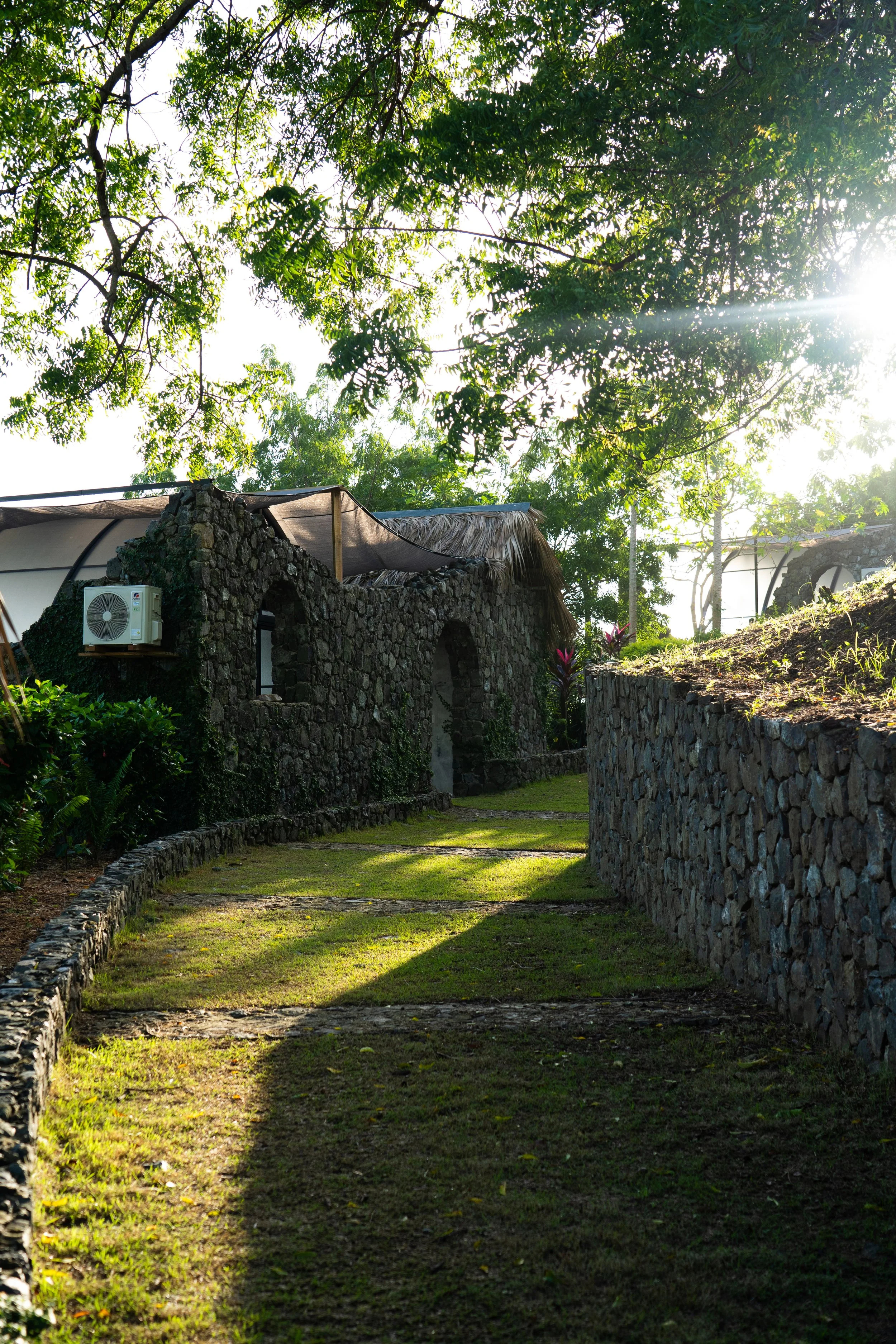 Stone pathway leading to rustic stone buildings with thatched roofs, surrounded by lush greenery and trees, with sunlight shining through.