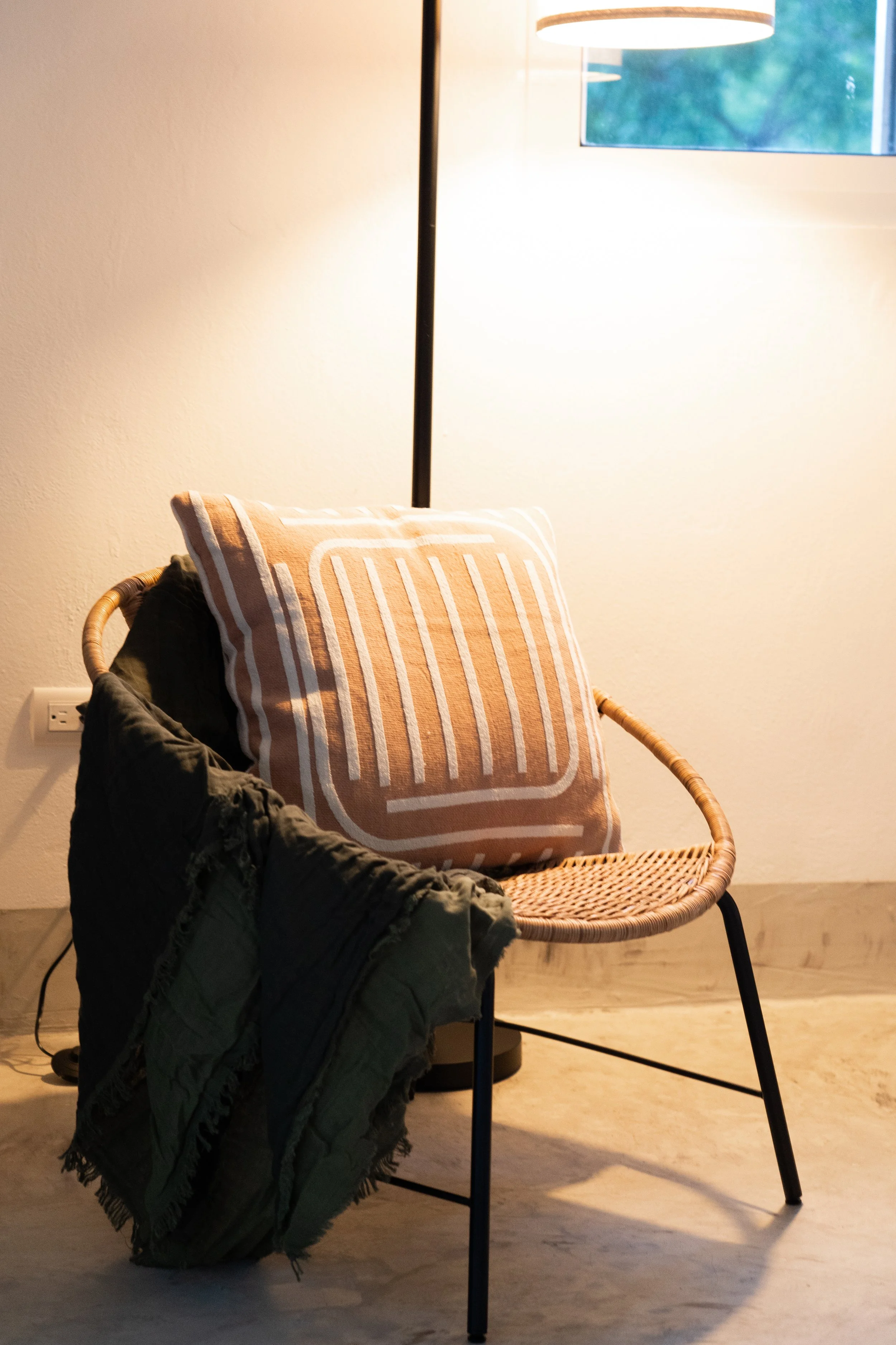 A wicker chair with a brown striped pillow and a dark green throw blanket draped over the side, positioned next to a black floor lamp near a window with blue sky and green trees visible outside.