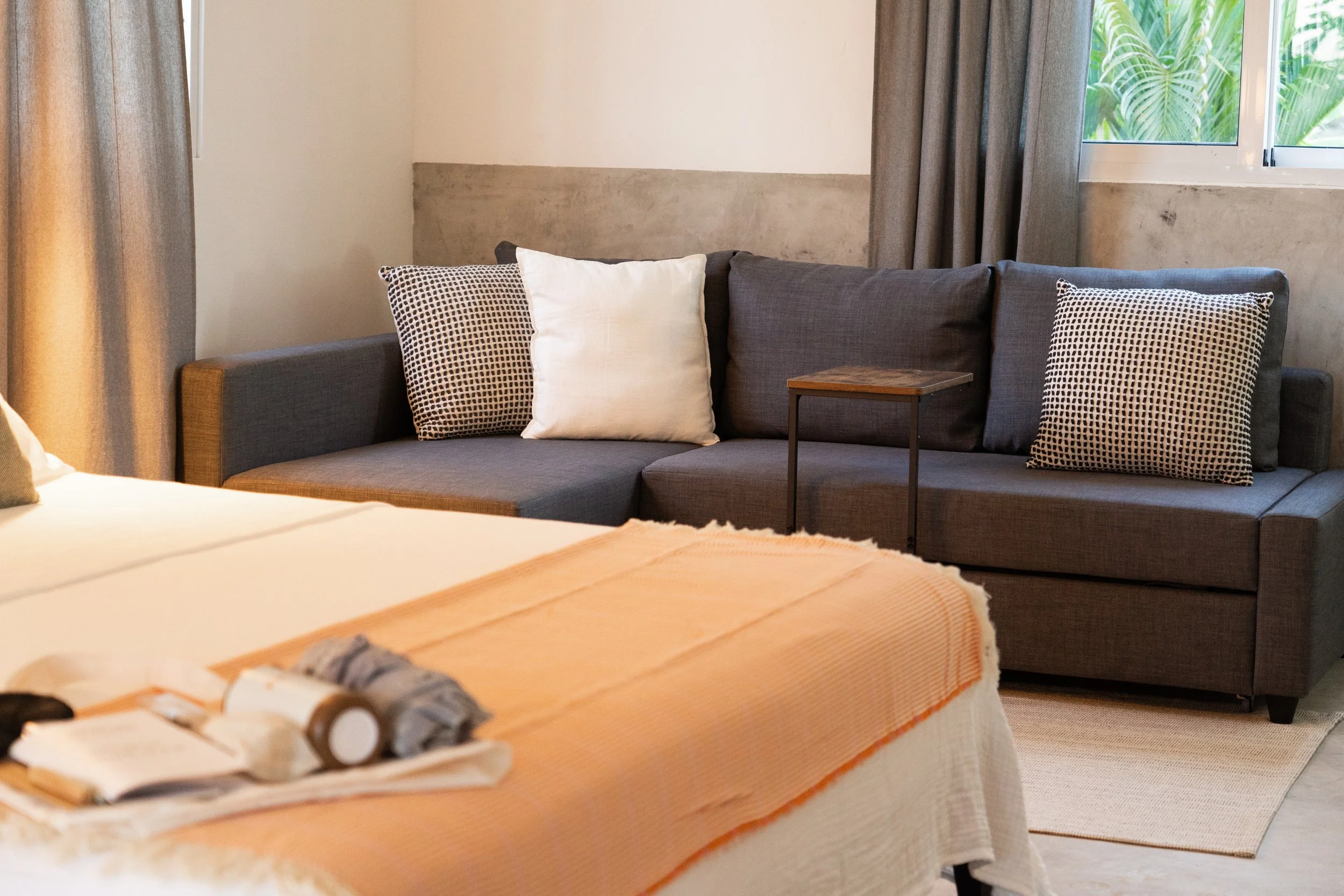 Hotel room with a bed in the foreground and a gray sofa with pillows near the window in the background.