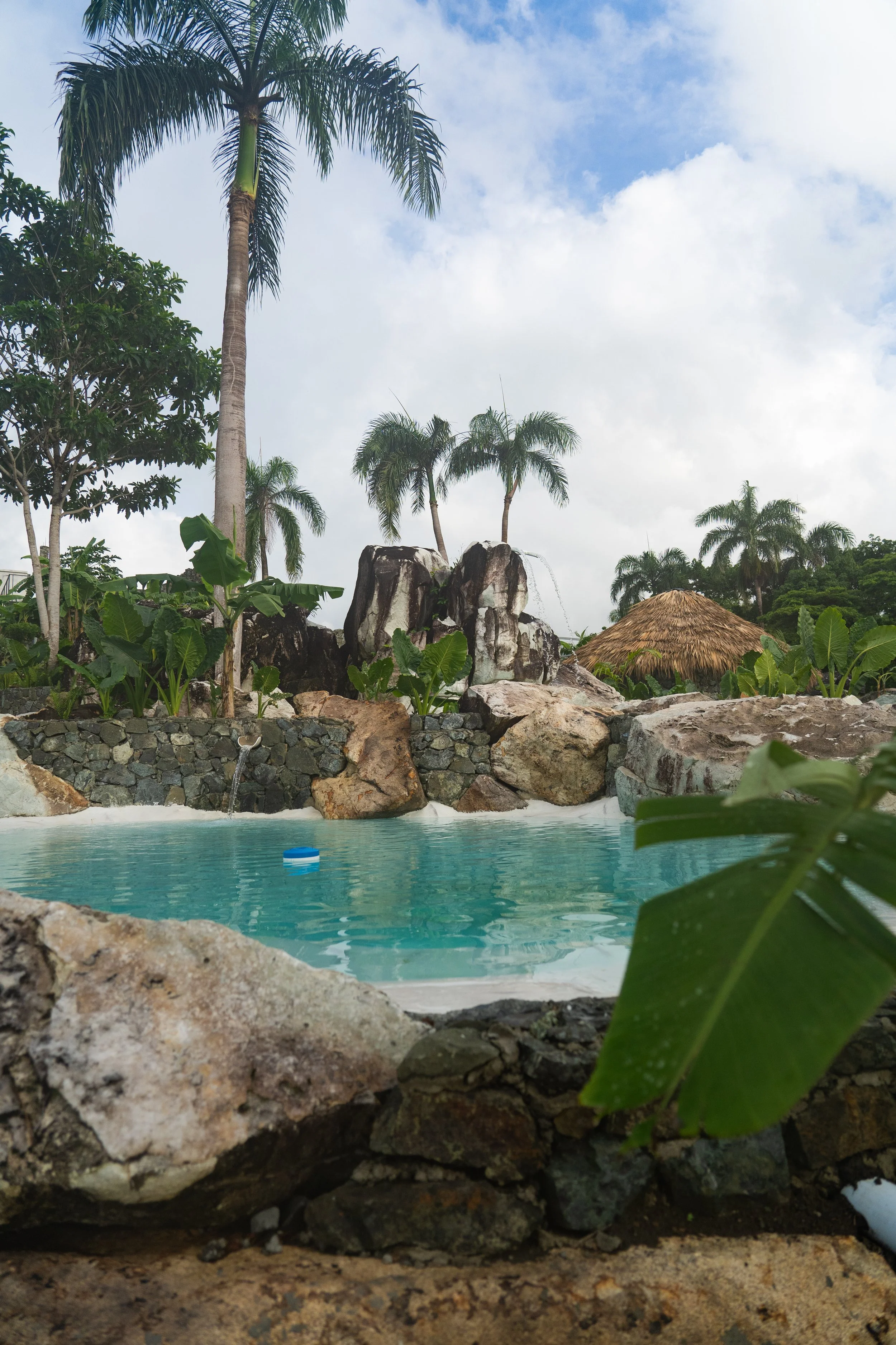 A tropical outdoor scene featuring a swimming pool surrounded by rocks, lush greenery, palm trees, and a thatched-roof hut under a partly cloudy sky.