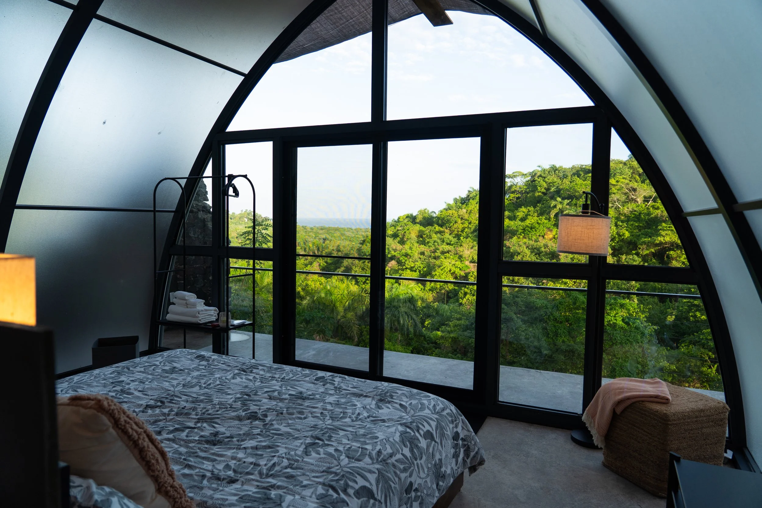 Interior of a modern bedroom with large arched windows overlooking lush green trees and hills, featuring a bed with patterned bedding, a lamp, and a wicker stool with a pink blanket.