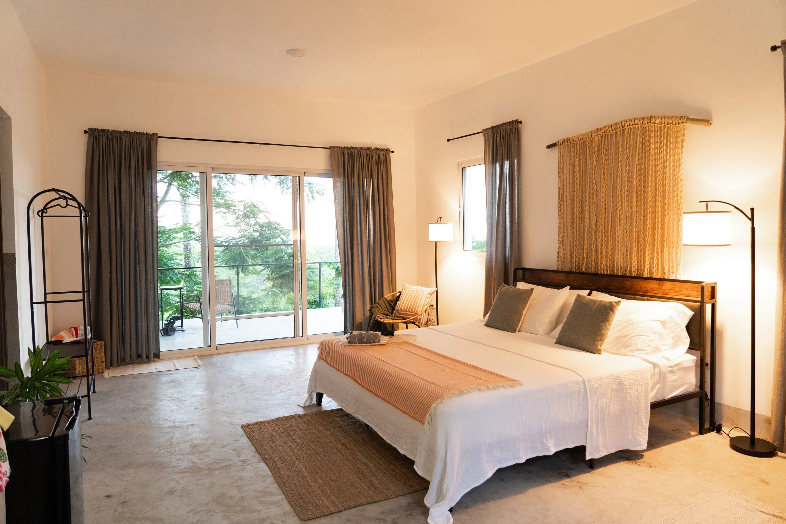 Bright hotel bedroom with king-sized bed, white linens, gray pillows, beige throw blanket, lighting fixtures, sliding glass door leading to a balcony with outdoor seating, and tropical greenery outside.