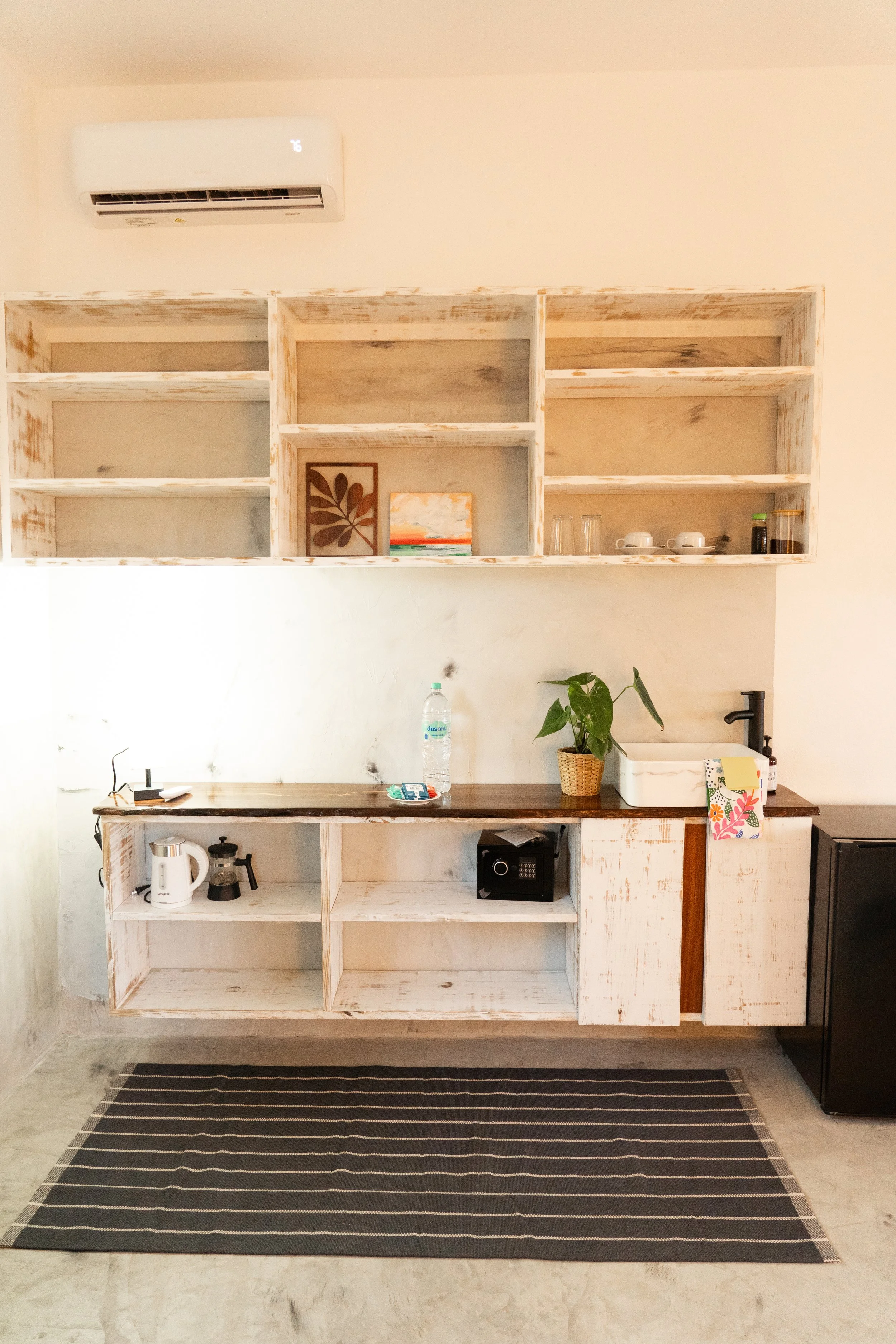 A minimalist kitchen with open wooden shelves, a countertop with a water bottle, a plant, a small safe, a kettle, and coffee-making equipment, set against a white wall with an air conditioning unit.