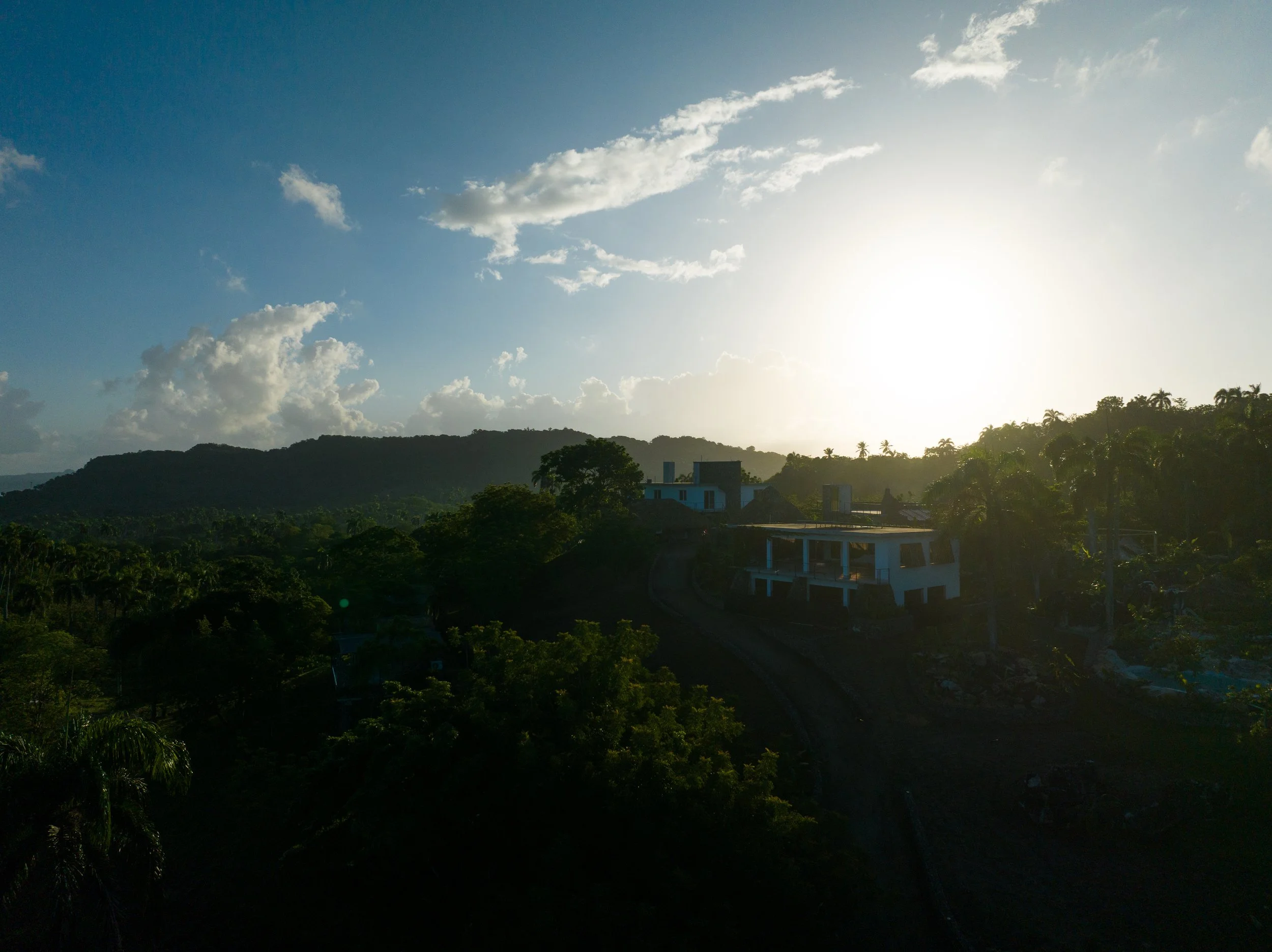 Mountains, trees, and houses on a hillside during sunset or sunrise with a bright sky and scattered clouds.