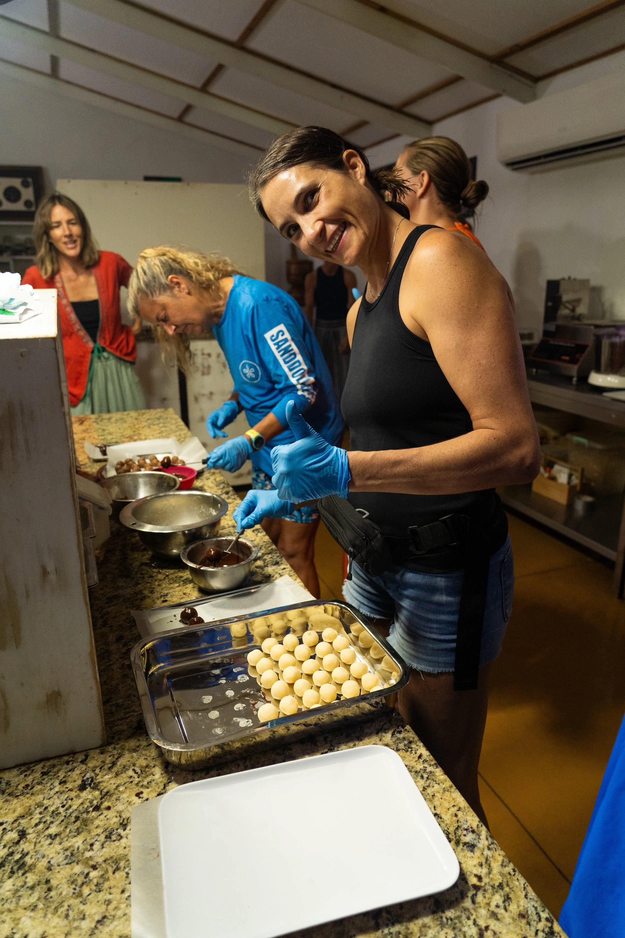 A group of women in a kitchen preparing chocolate bonbons, with one woman smiling at the camera and wearing blue gloves.