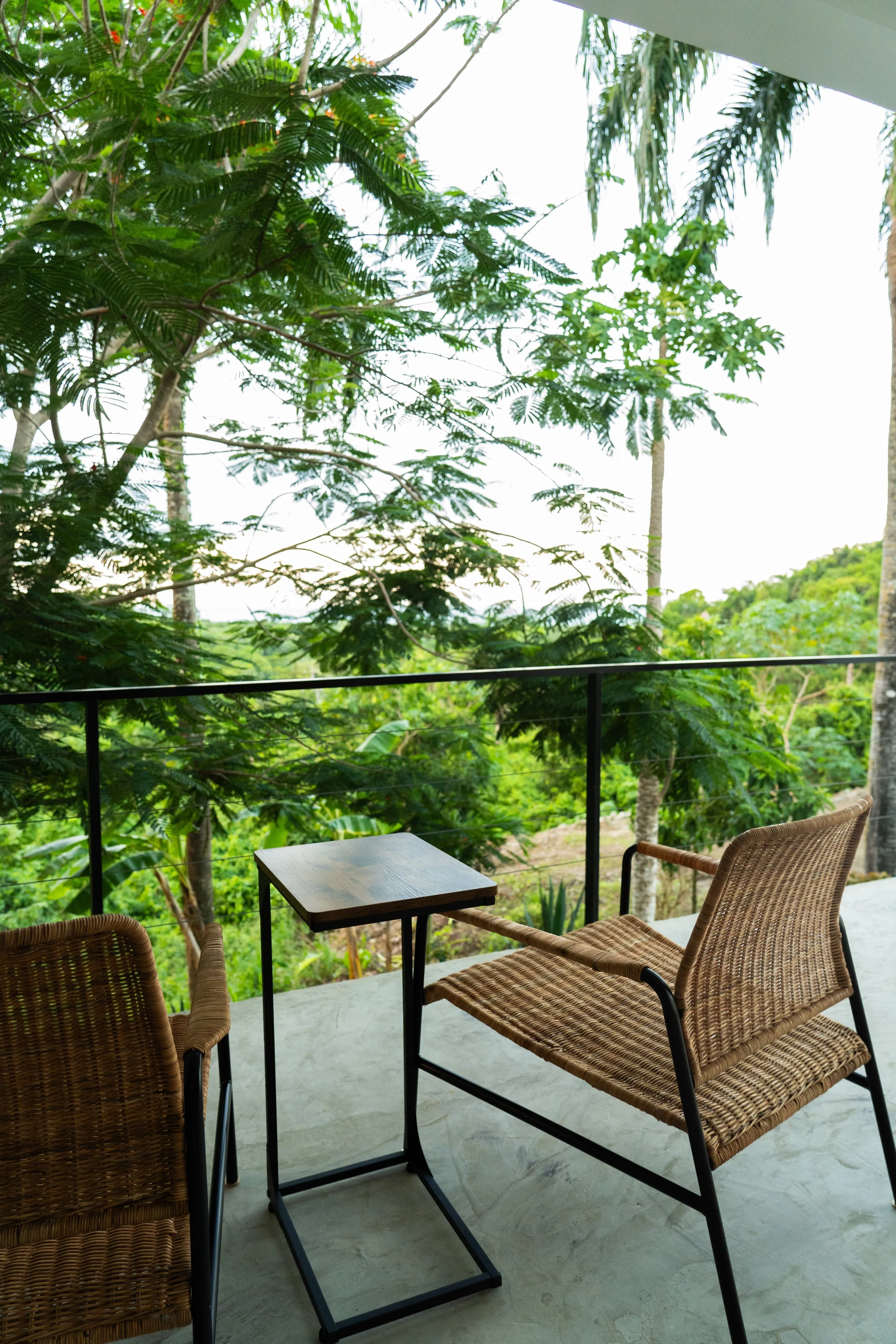 Balcony with two wicker chairs, small wooden table, overlooking lush green forest with tall trees and jungle foliage.
