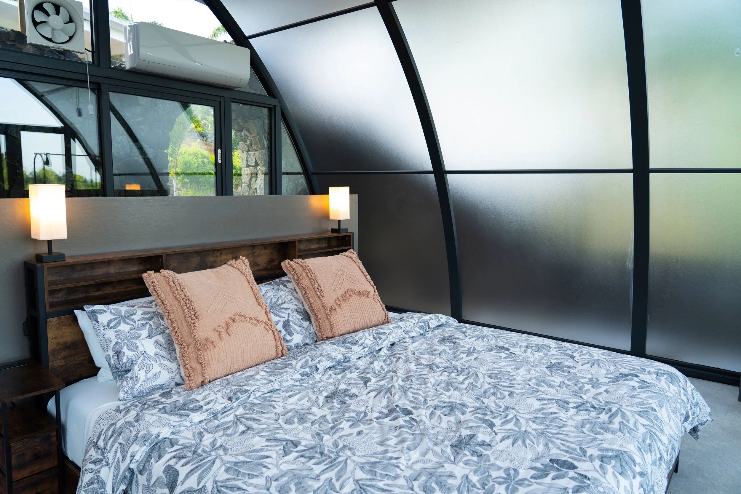 A bedroom with a large bed featuring floral bedding, two beige pillows, and a wooden headboard flanked by wall-mounted lamps, inside a room with large glass windows and sliding doors, and a frosted glass wall on the right.