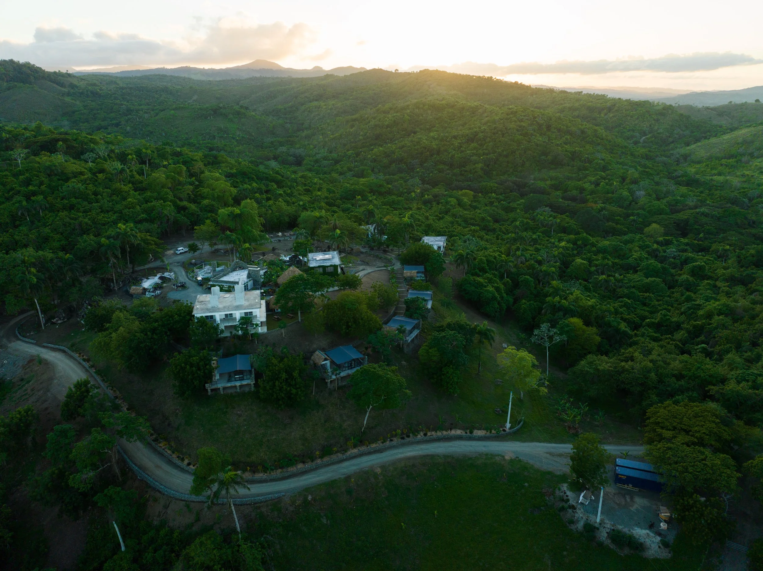 Aerial view of a small residential area surrounded by dense green forest and mountains, with a winding dirt road leading to the houses on a hillside at sunset.