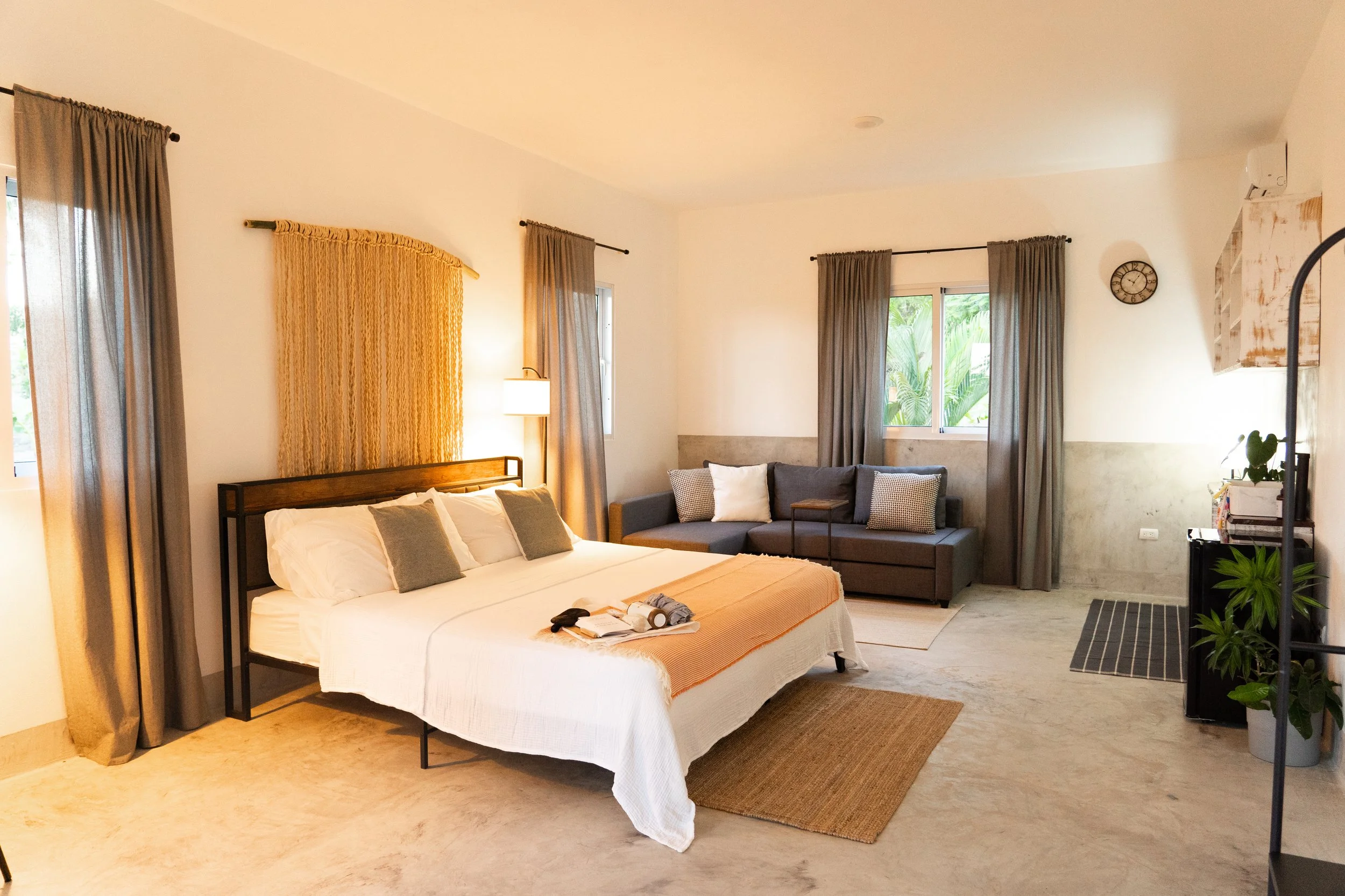 A spacious bedroom with a large bed, a gray sofa with cushions, multiple windows with curtains, a small side table, indoor plants, and neutral-colored decor.