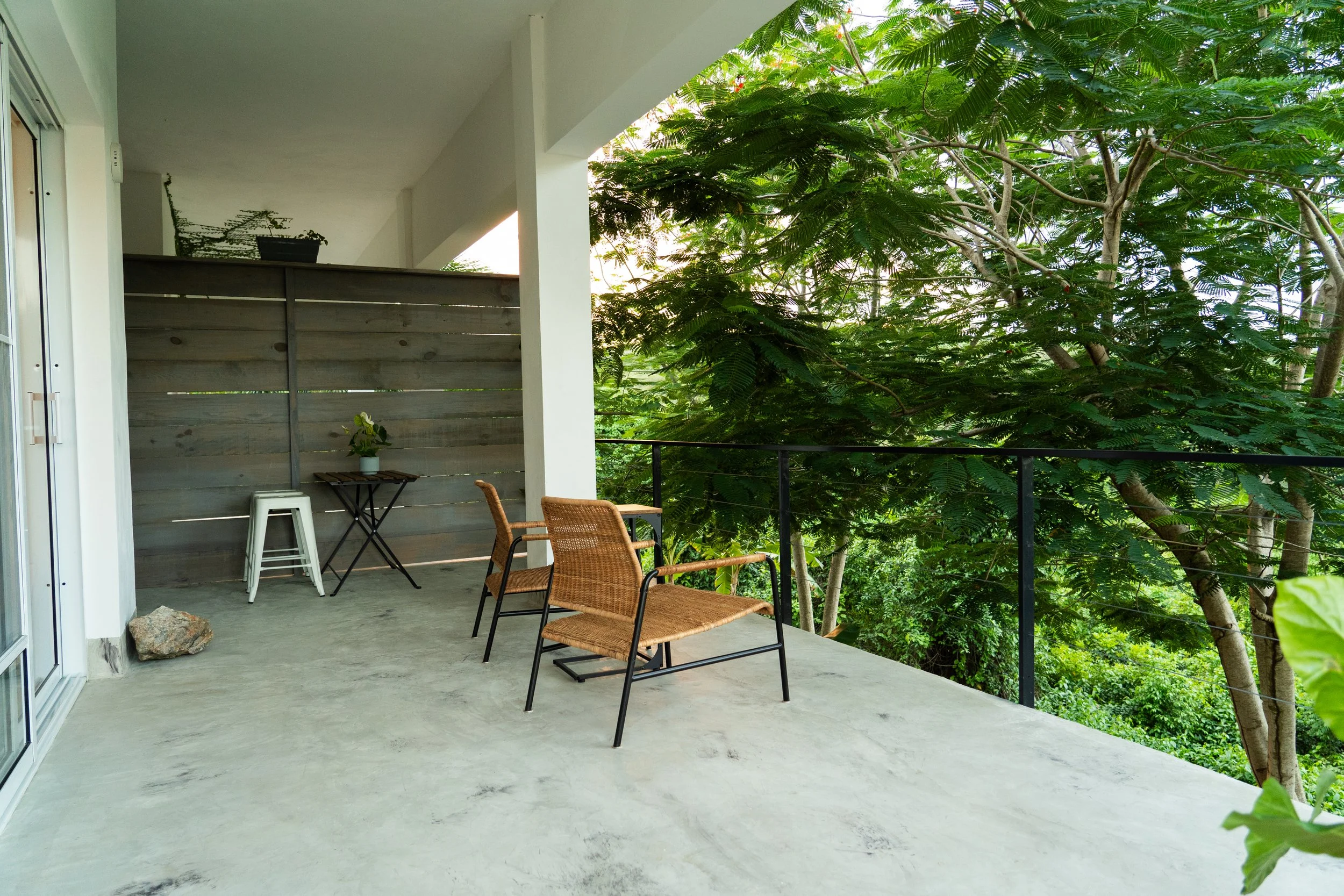 Balcony with two rattan chairs, a small table with a plant, and a wooden privacy fence, overlooking lush green trees.