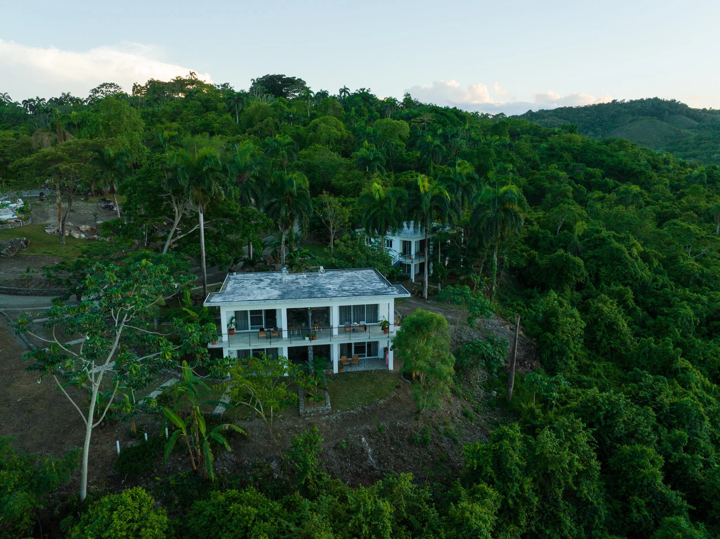 An aerial view of a house on a hillside surrounded by dense green forest and mountains in the background.