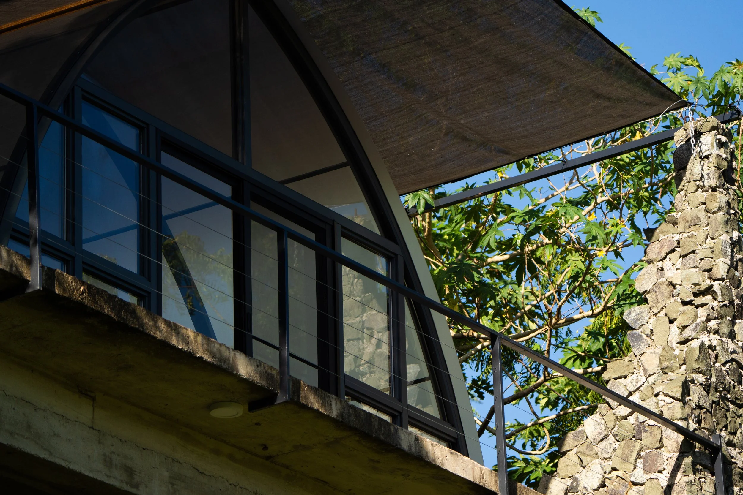 Close-up view of a modern house with glass windows, a concrete balcony, a metal railing, a stone chimney, and trees in the background under a clear blue sky.