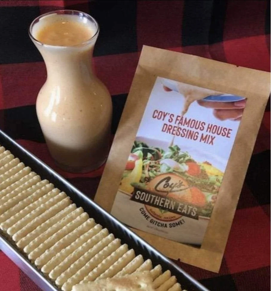 A bottle of creamy, Coy's dressing, a signature tray of saltine crackers, and a packet labeled 'Coy's Famous House Dressing Mix' on a checkered table.