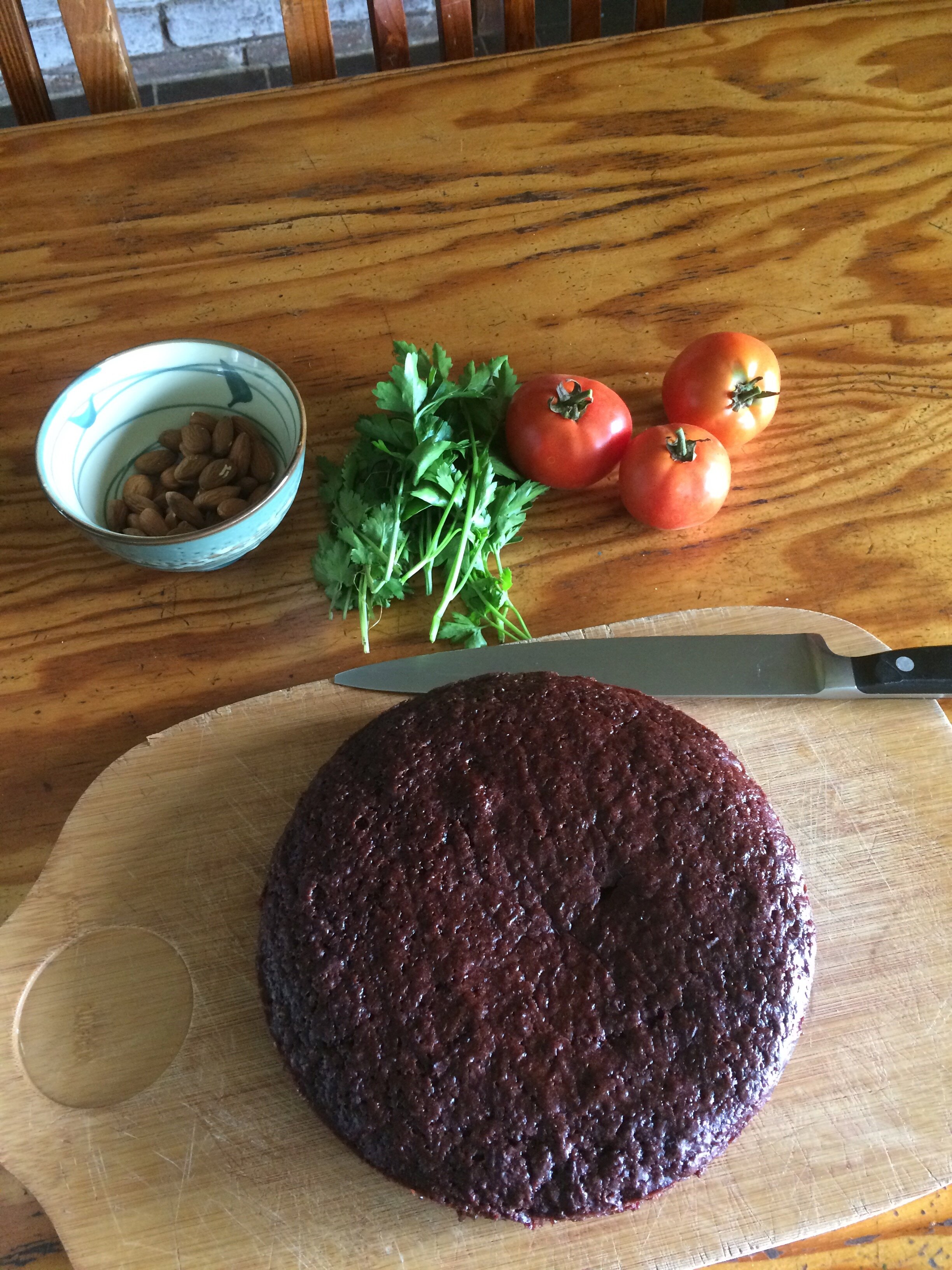 Top view of round brown cake on chopping board next to basil, tomatoes, bowl of nuts