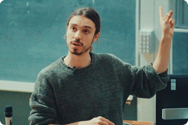 Young white man with dark goatee and knitted jumper pointing up