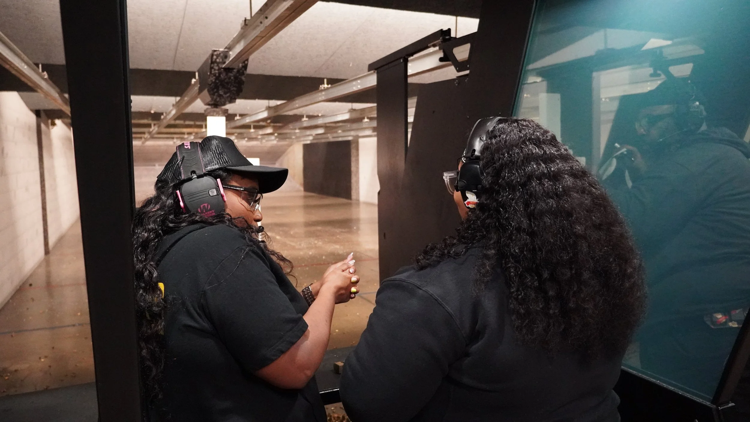 Two women at an indoor shooting range, one is holding a small item and the other is looking at it. Both are wearing black shirts, safety glasses, and hearing protection.