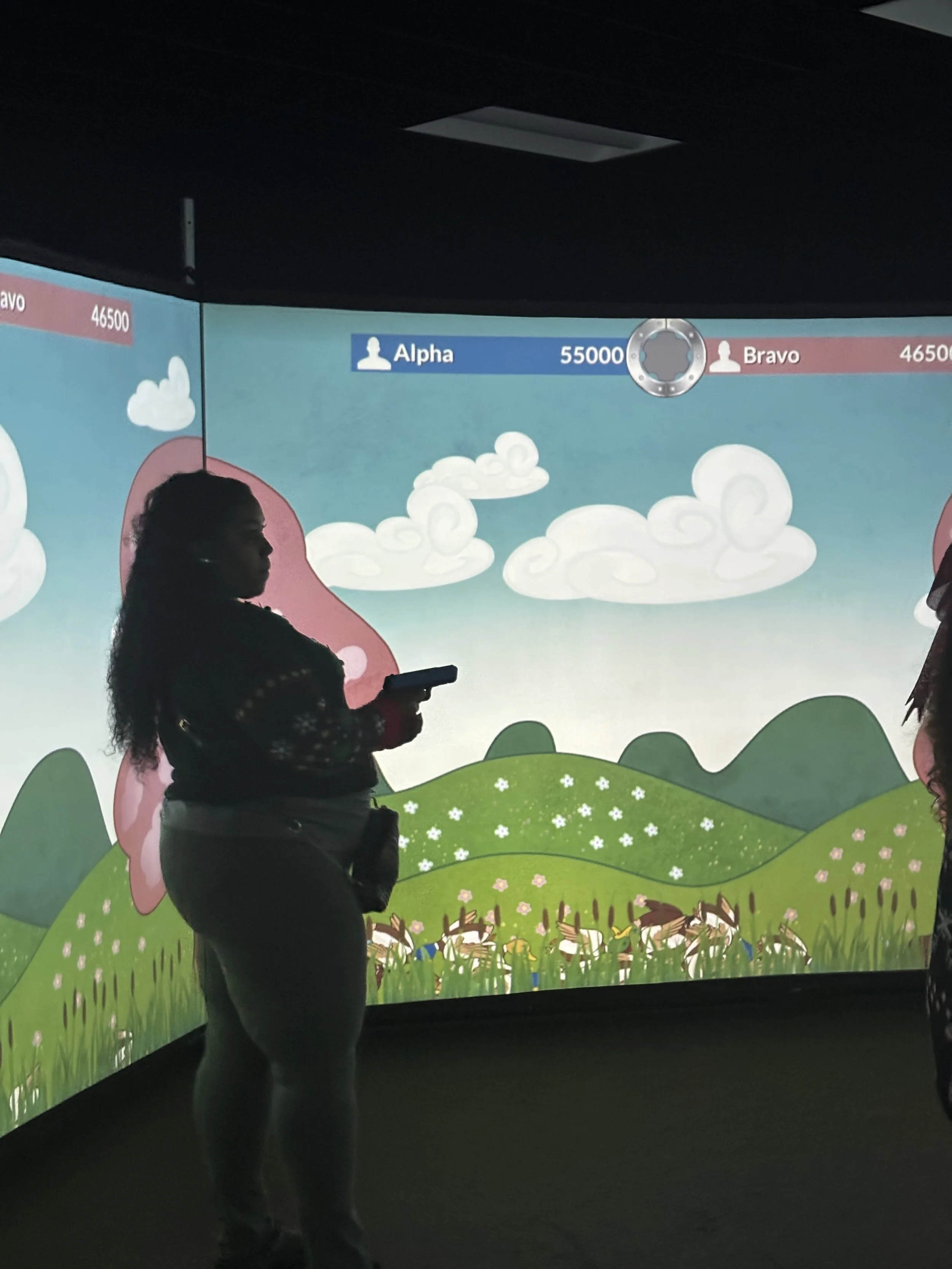 Silhouette of a woman holding a phone in front of a large screen displaying a digital game with mountains, clouds, and information about two teams, Alpha and Bravo, with scores 55,000 and 46,500.