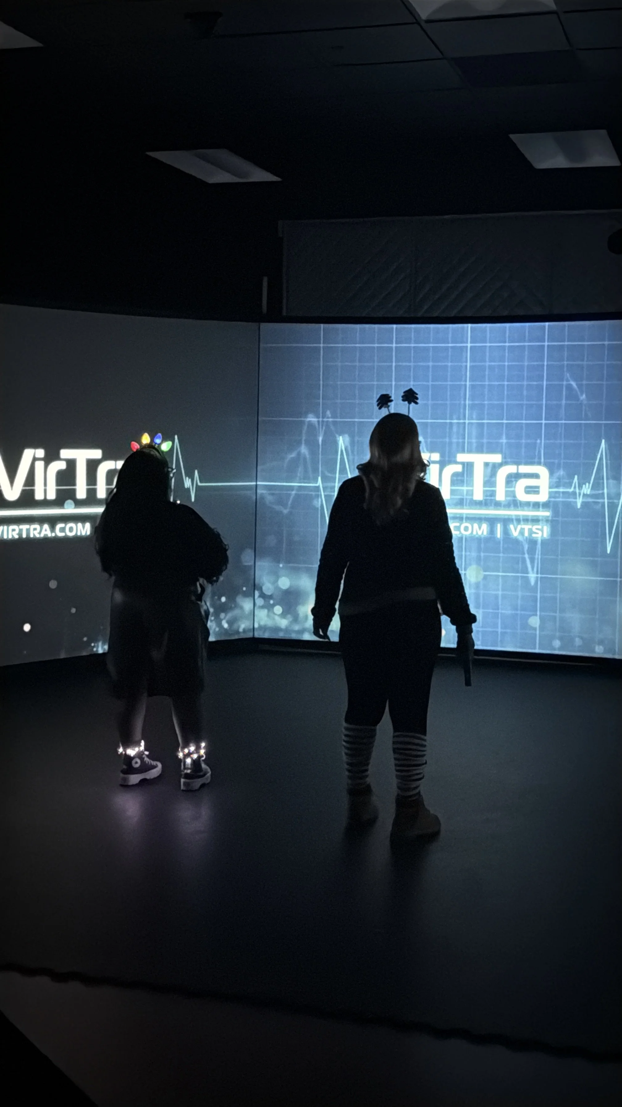 Silhouettes of two children standing in front of a large digital screen displaying a heartbeat line, the website 'VIRTRA.COM', and the text 'VIRTRA', in a dark room.