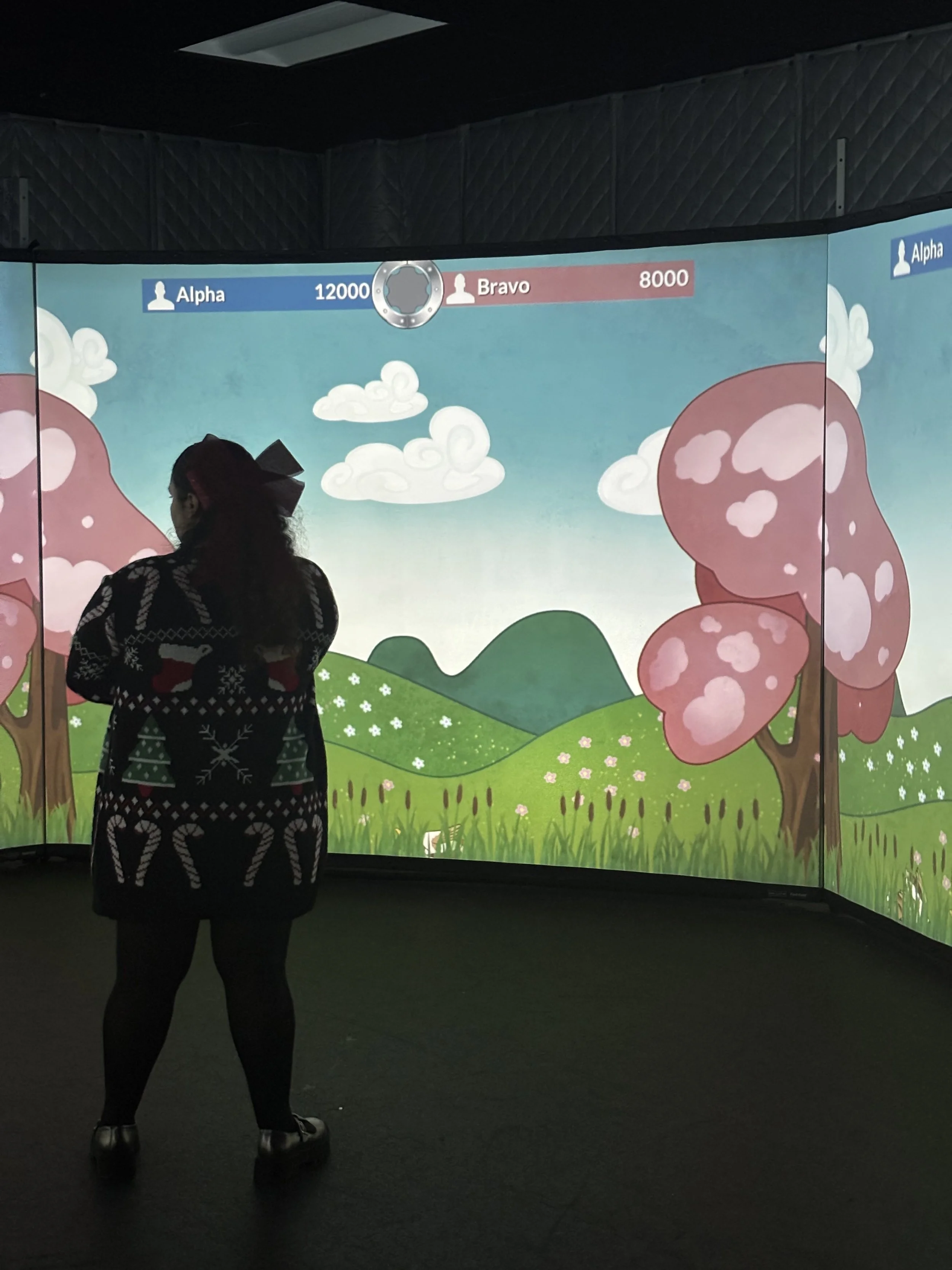 A person standing in front of a large video game screen with animated scenery of trees and hills, showing scores for teams Alpha and Bravo.