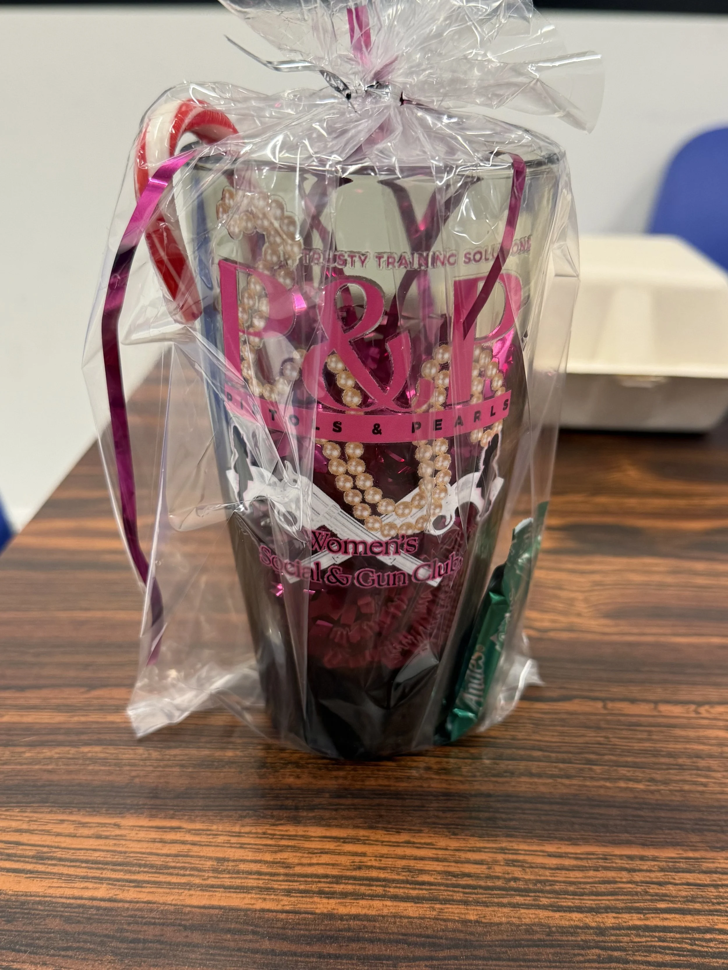 A cellphone wrapped in clear plastic with pink lettering that reads 'P&R' and 'Women's Social & Gun Club,' placed on a wooden table. The phone case features pearls and other decorative elements.