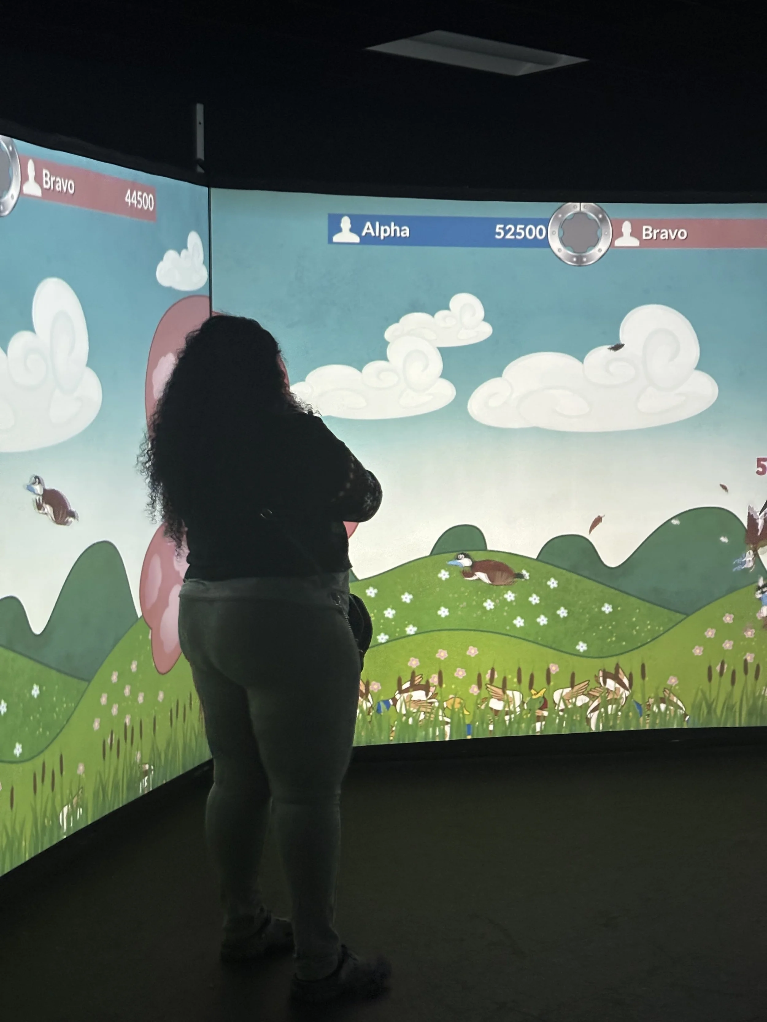 Person standing in front of a large, curved, colorful gaming screen with cartoonish landscape and statistics for two teams, Alpha and Bravo.