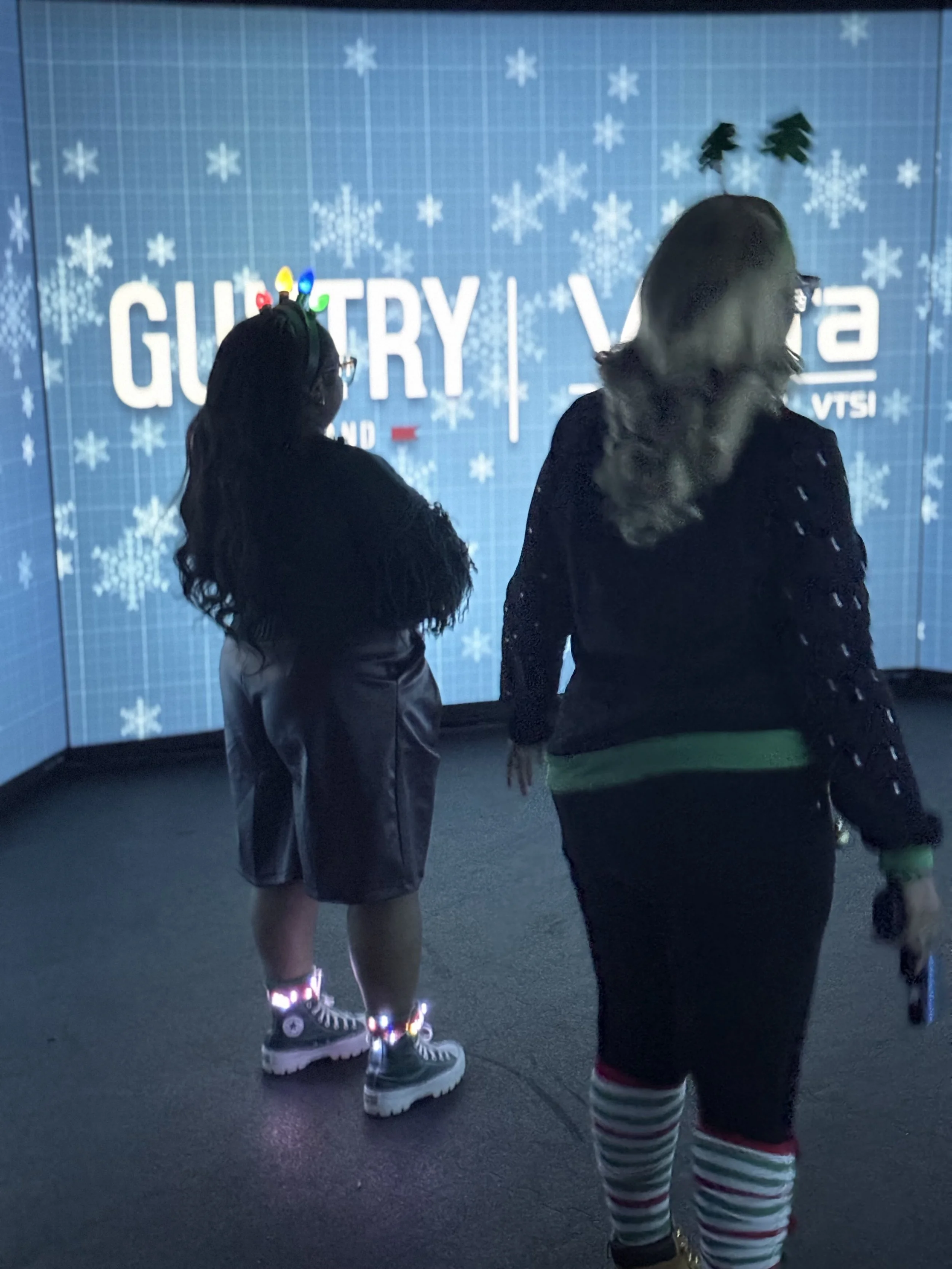 Two people standing in front of a blue wall with snowflake patterns and text, one wearing reindeer antlers and colorful Christmas lights on their head, the other with reindeer antlers and striped stockings, both facing away from the camera.