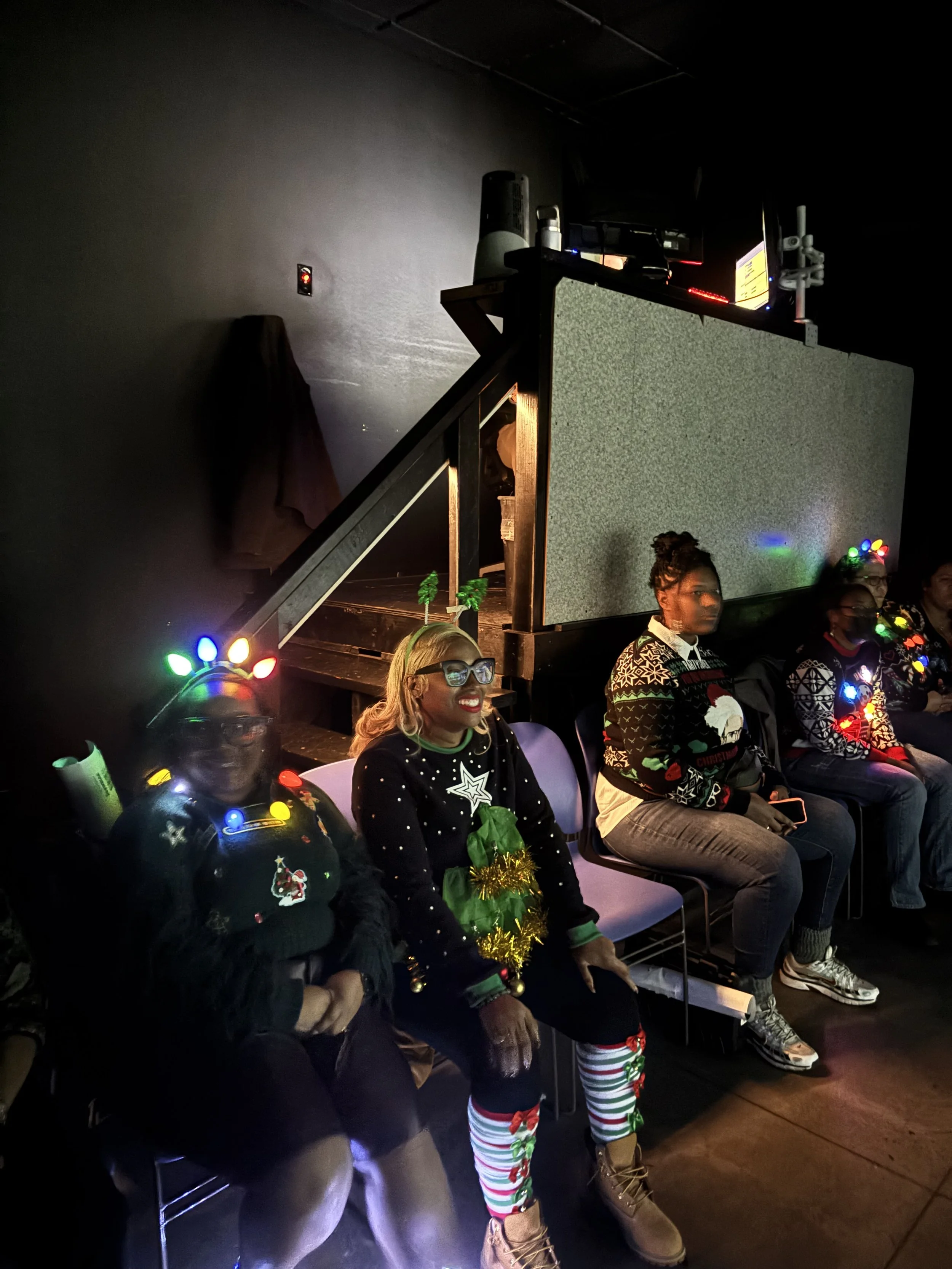 People sitting in a row at a holiday event, wearing Christmas sweaters and accessories, with colorful lights and decorations.