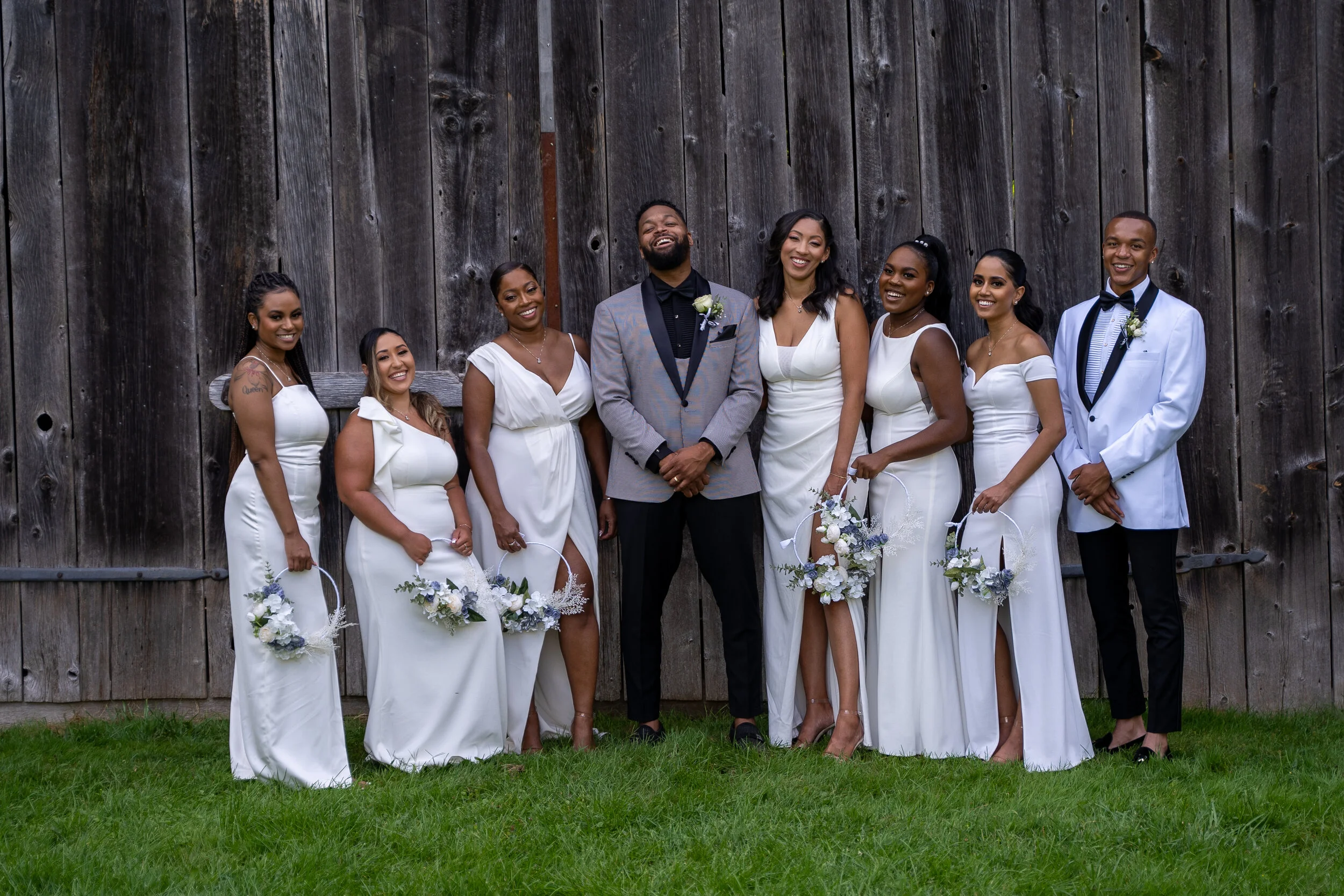 Group of wedding guests dressed in white, standing outdoors in front of a wooden fence, smiling and posing for the photo.