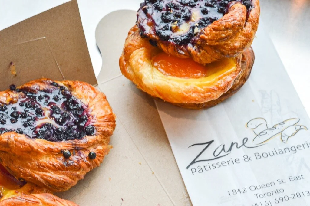 Three blueberry Danish pastries with flaky golden crusts, some featuring a peach filling, on a Zane Pâtisserie & Boulangérie paper and cardboard box.