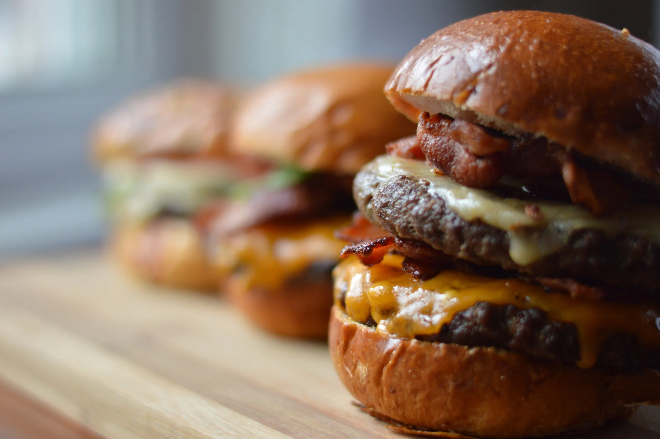 Close-up of three beef cheeseburgers on a wooden surface, showing buns, cheese, beef patties, bacon, and other ingredients.