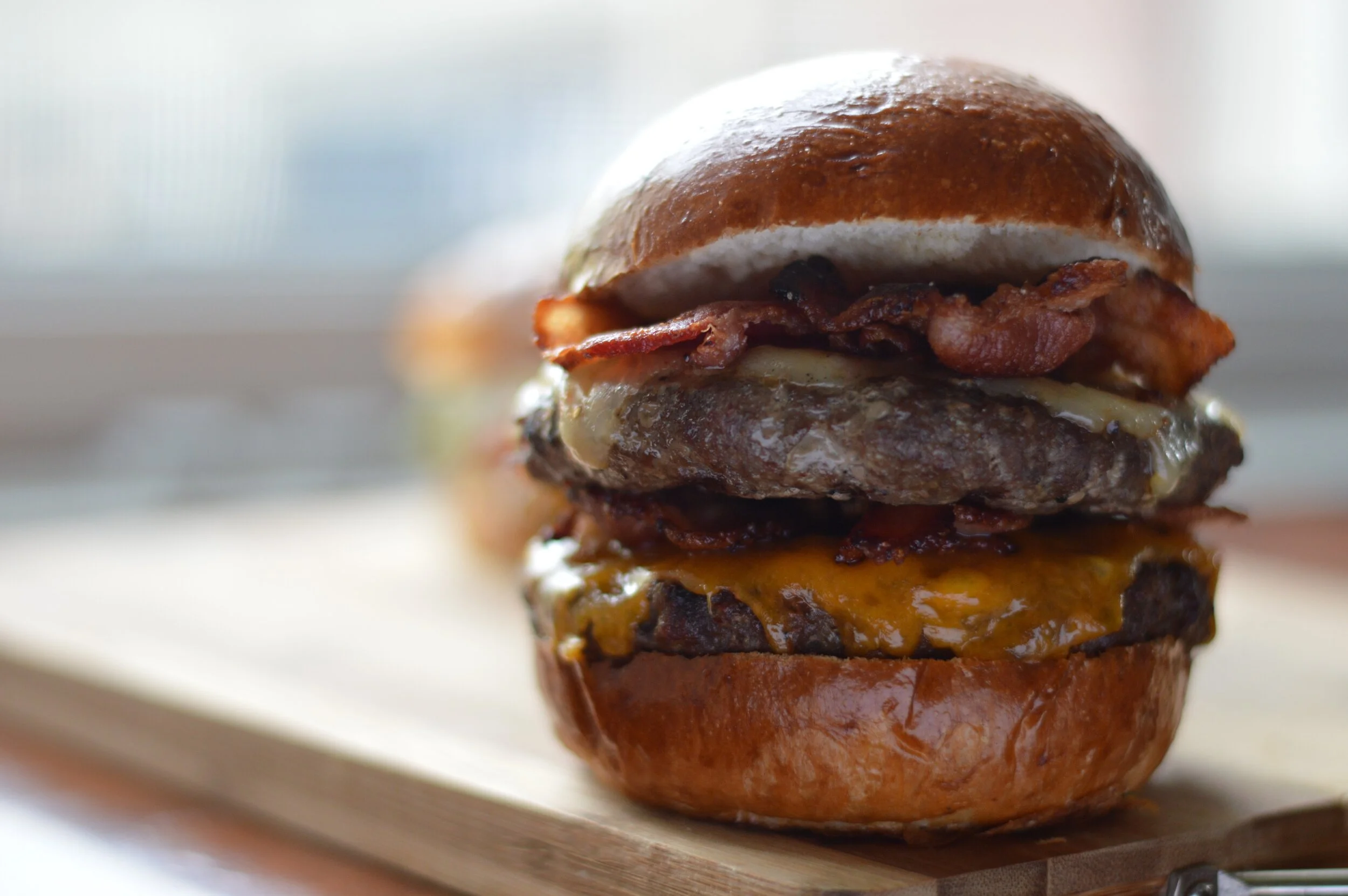 Close-up of a multi-layer cheeseburger with bacon, melted cheese, and beef patty on a wooden surface.