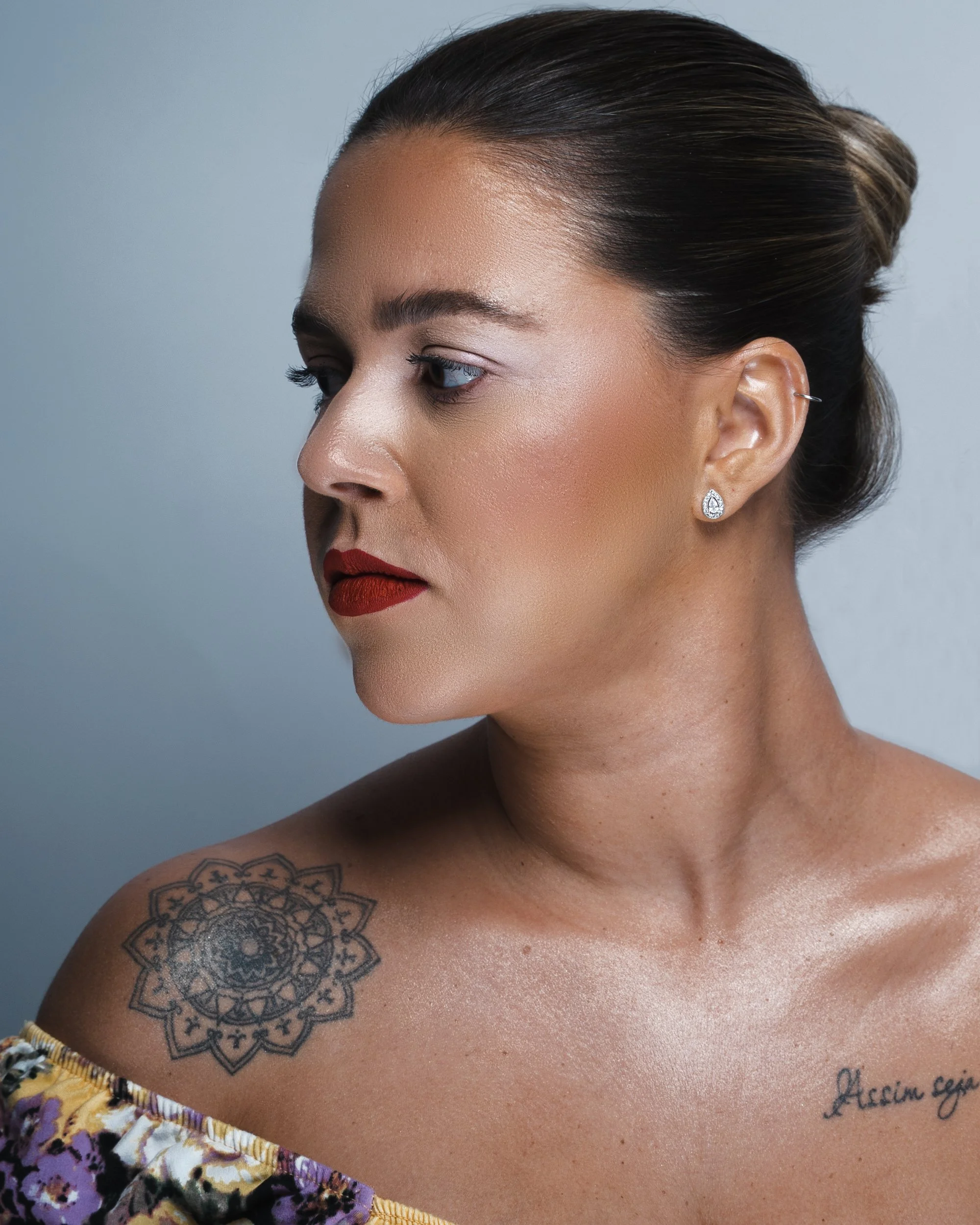 Side profile of a woman with dark hair in a bun, red lipstick, and tattoo on her shoulder, wearing earrings and an off-shoulder floral top.