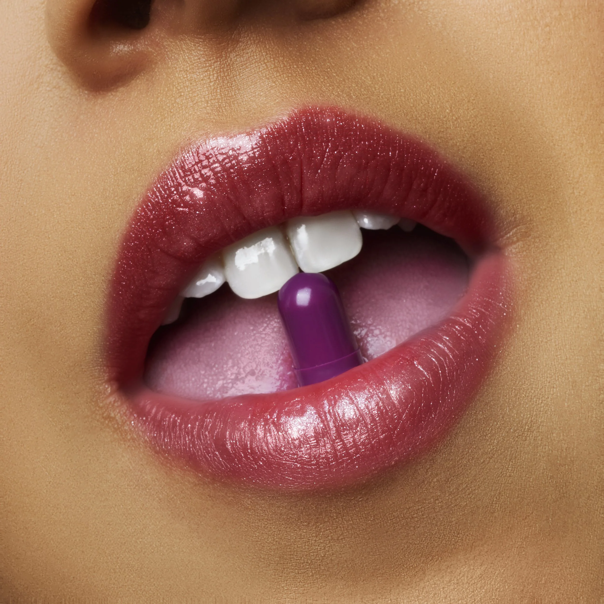 Close-up of a person's lips with glossy pink lipstick, holding a purple capsule pill between their teeth, with part of their teeth and tongue visible.
