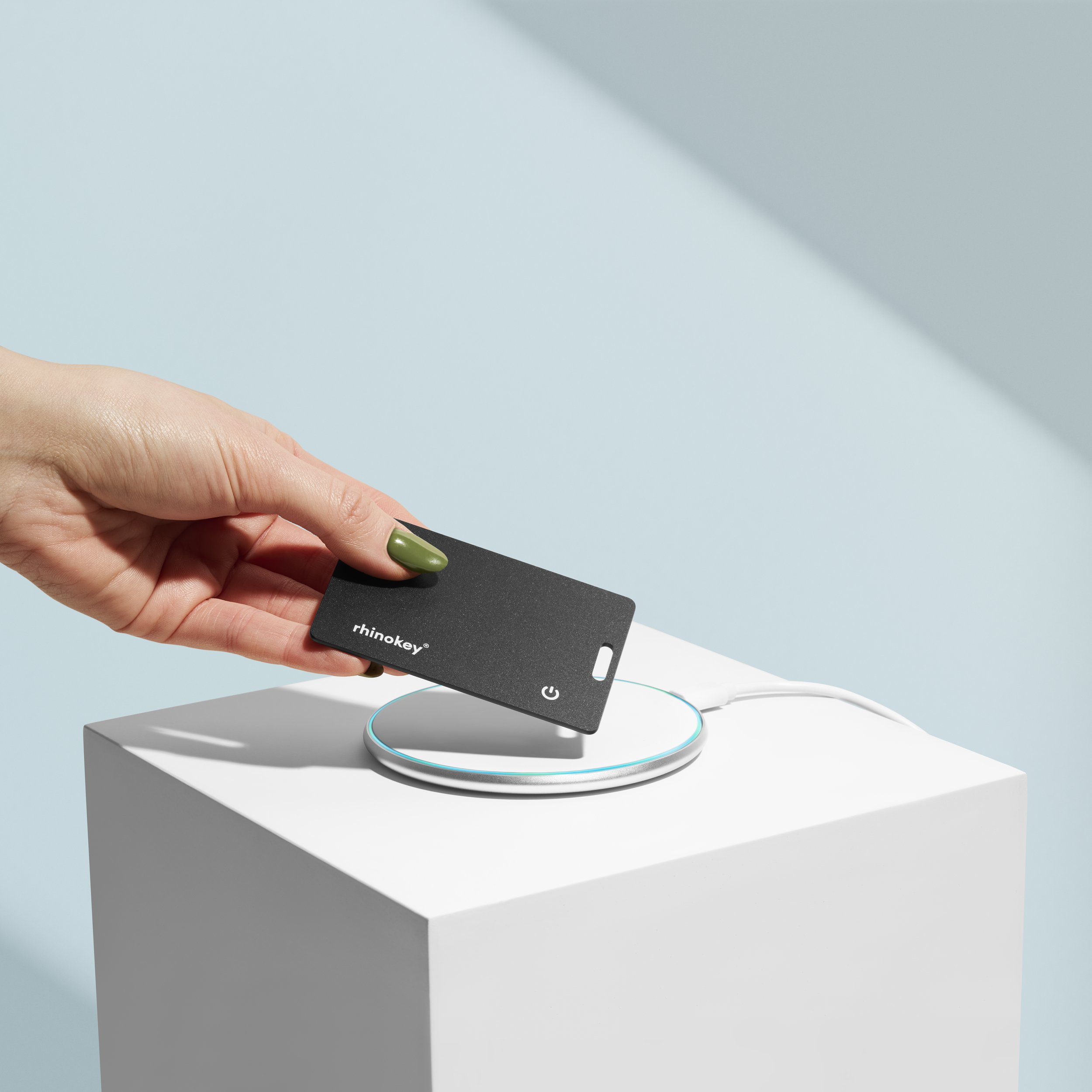 A hand with green nails holding a black RFID card labeled 'rhinokey' above a white wireless charging pad. Product Photography. Creative Photography. eCommerce Photography.