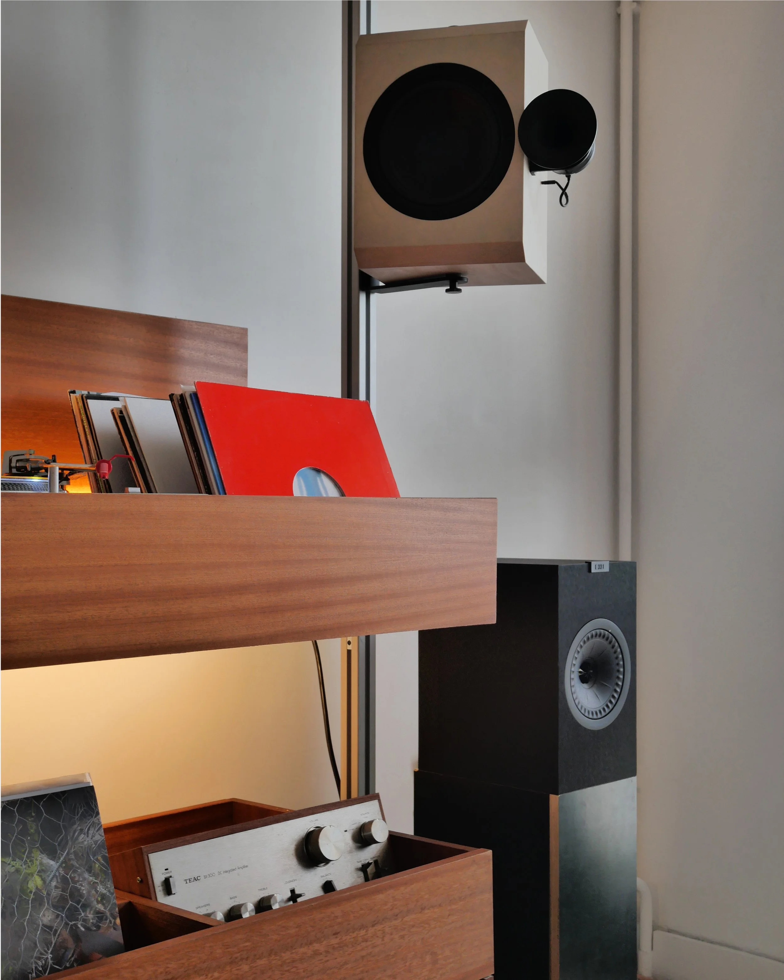 Close-up view of a large black speaker mounted on a wooden stand, with an amplifier in the foreground and a partial view of a book or magazine, along with some audio equipment and cables.