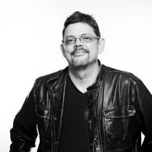 Black and white portrait of a man with glasses, beard, and short hair, wearing a leather jacket and smiling.