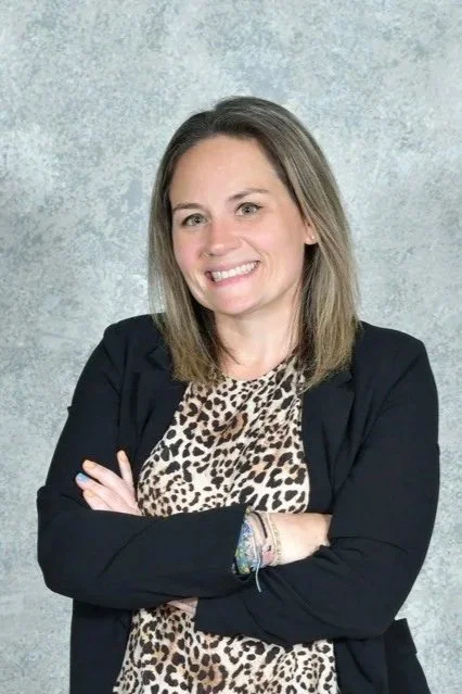 A woman with shoulder-length light brown hair smiling, wearing a black blazer and leopard print top, standing against a gray textured background.