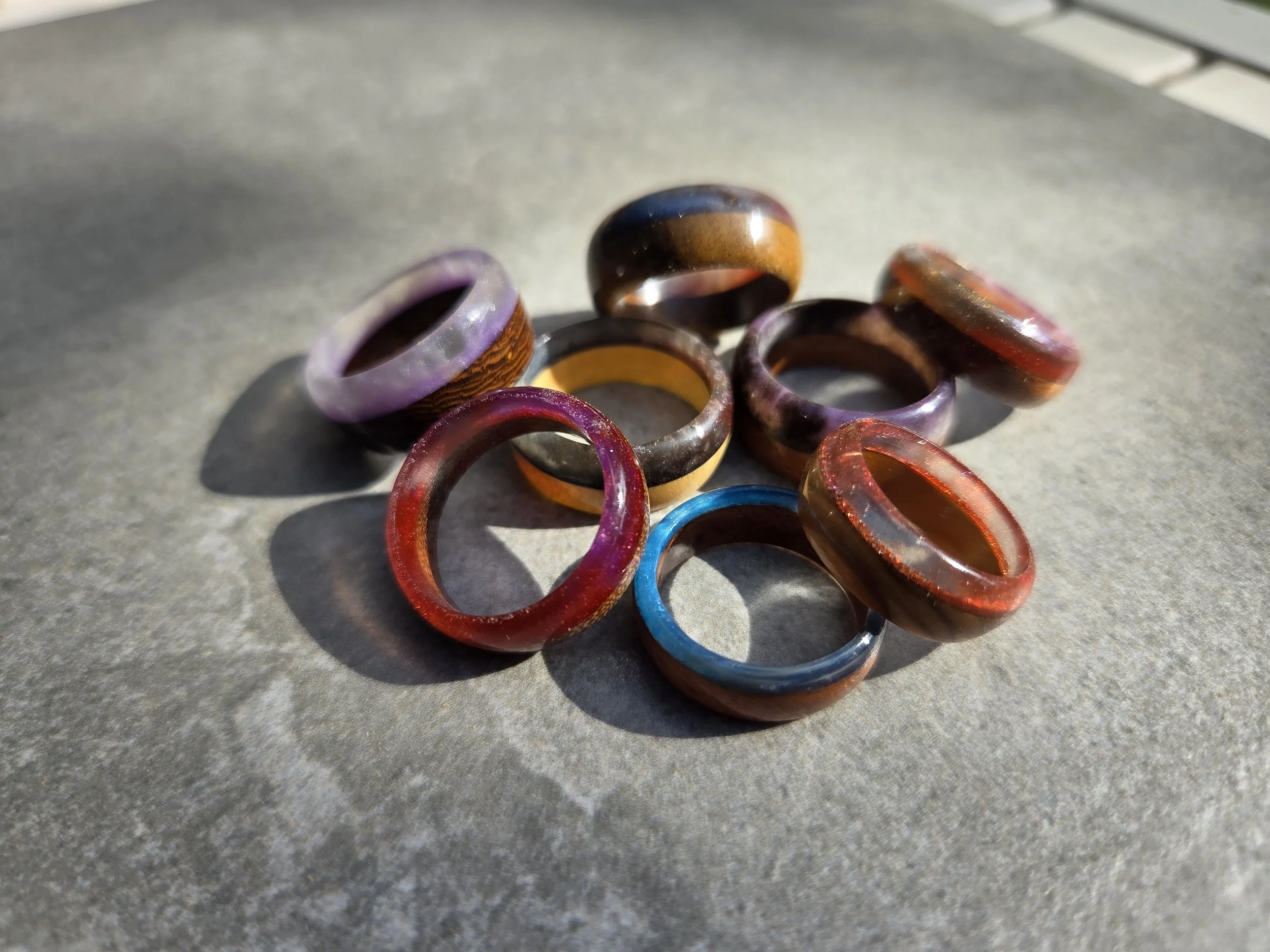 A collection of handmade wood and colourful resin rings by Rolf Designs on a grey stone surface.