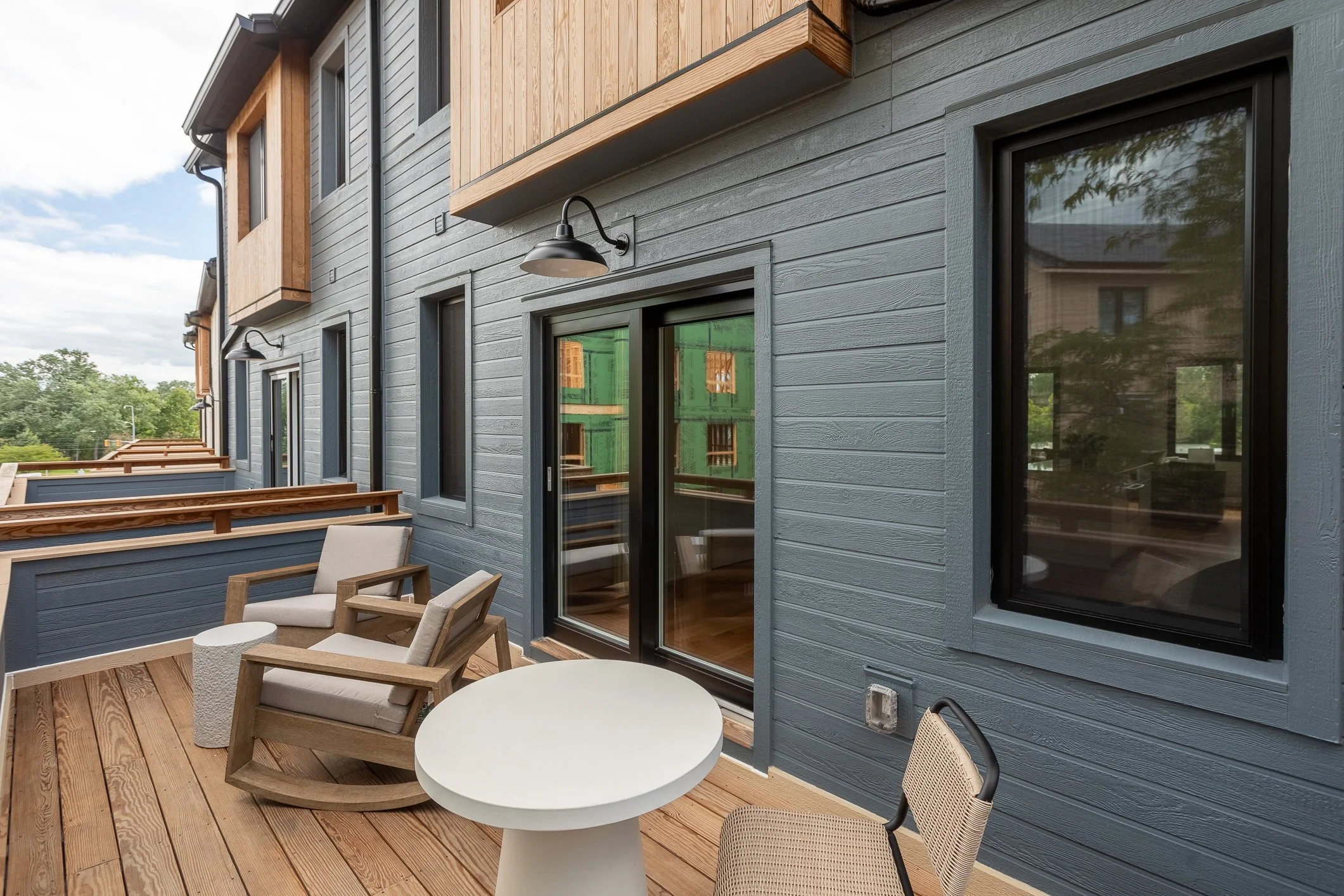 Outdoor balcony with wooden flooring, two chairs, and a round table, adjacent to a blue house with black-framed windows and sliding glass door.