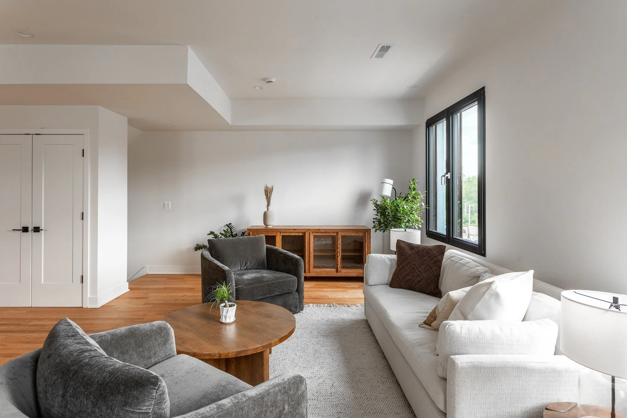 Living room with white walls, hardwood floor, large windows, white sofa with cushions, gray armchairs, a wooden coffee table with a small potted plant, a wooden console table with decorations, and various potted plants.