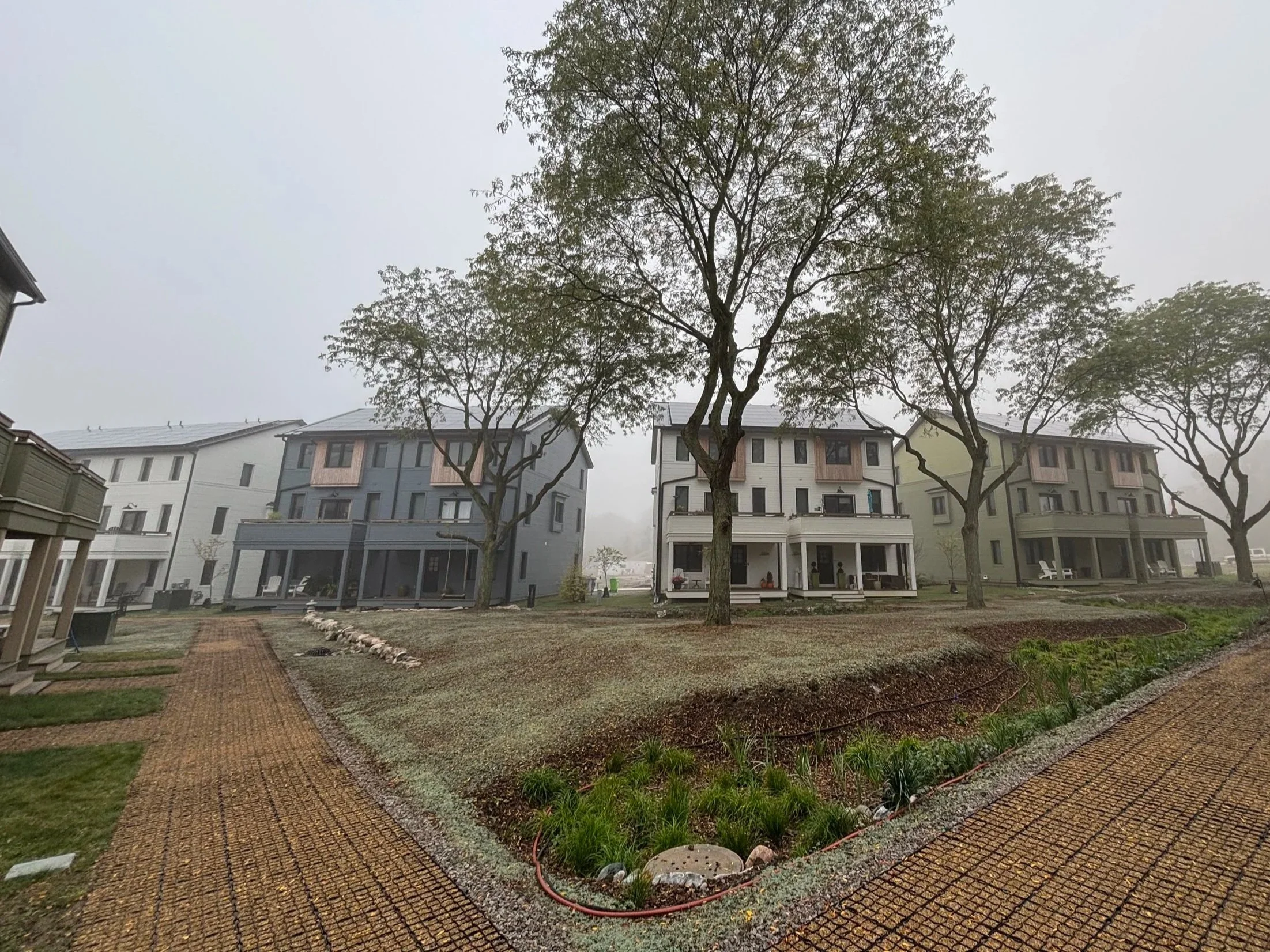 Multiple modern multi-story houses with porches and large windows, surrounded by trees and a landscaped yard with pathways and garden beds, under a foggy sky.
