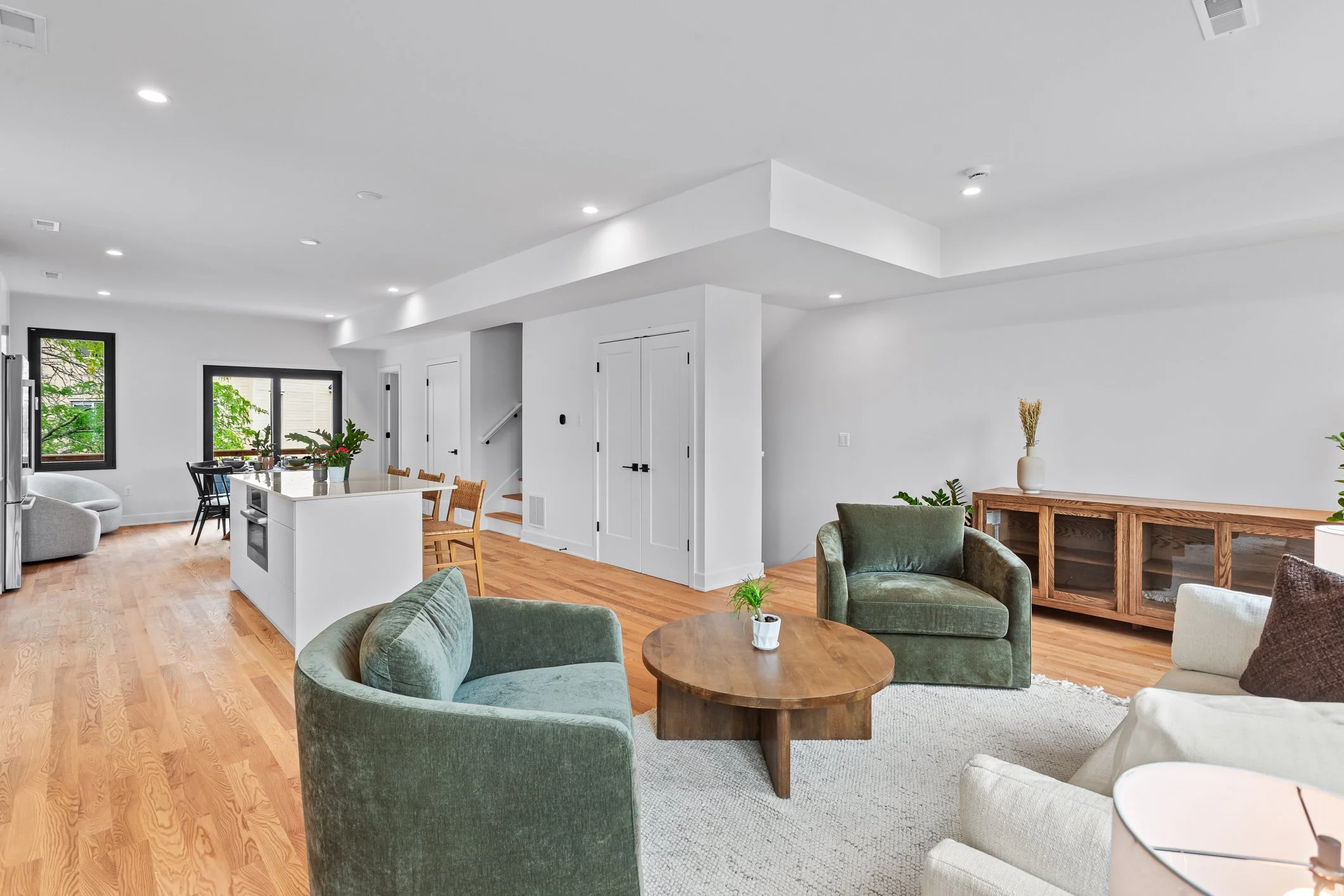 Open-concept living room and kitchen with white walls, wooden flooring, and modern furniture, including green and white chairs, a wooden coffee table, and a white kitchen island with barstools.