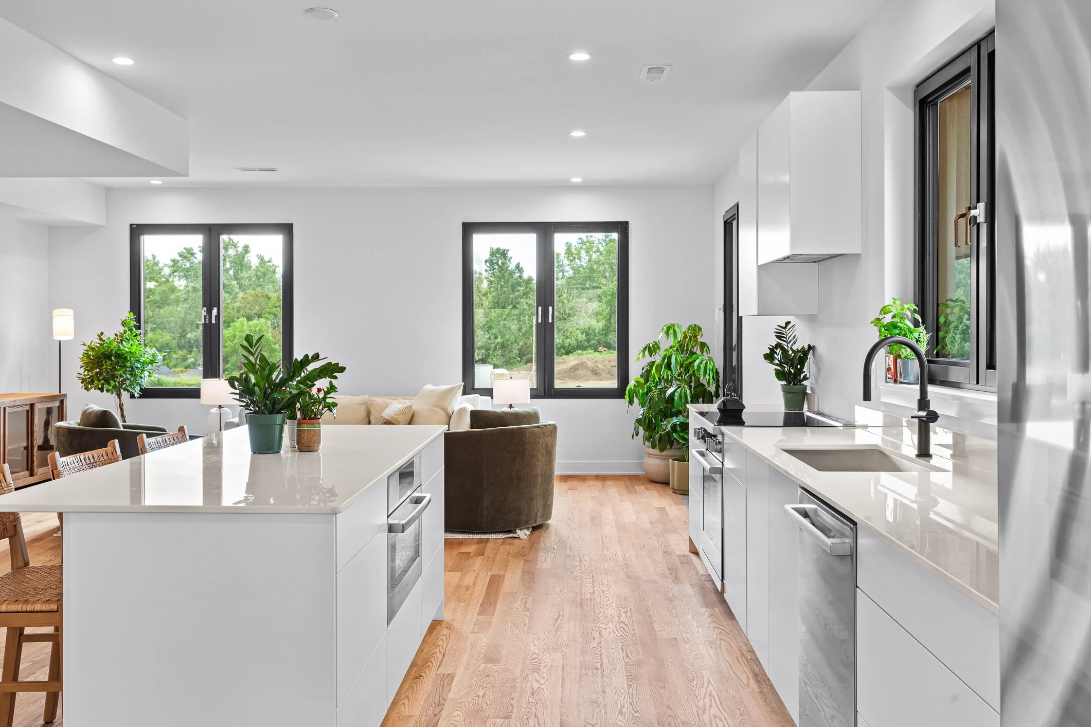 Modern open-concept kitchen with white cabinets, a large white island, and stainless steel appliances, overlooking a living area with beige sofas and large windows with greenery outside.