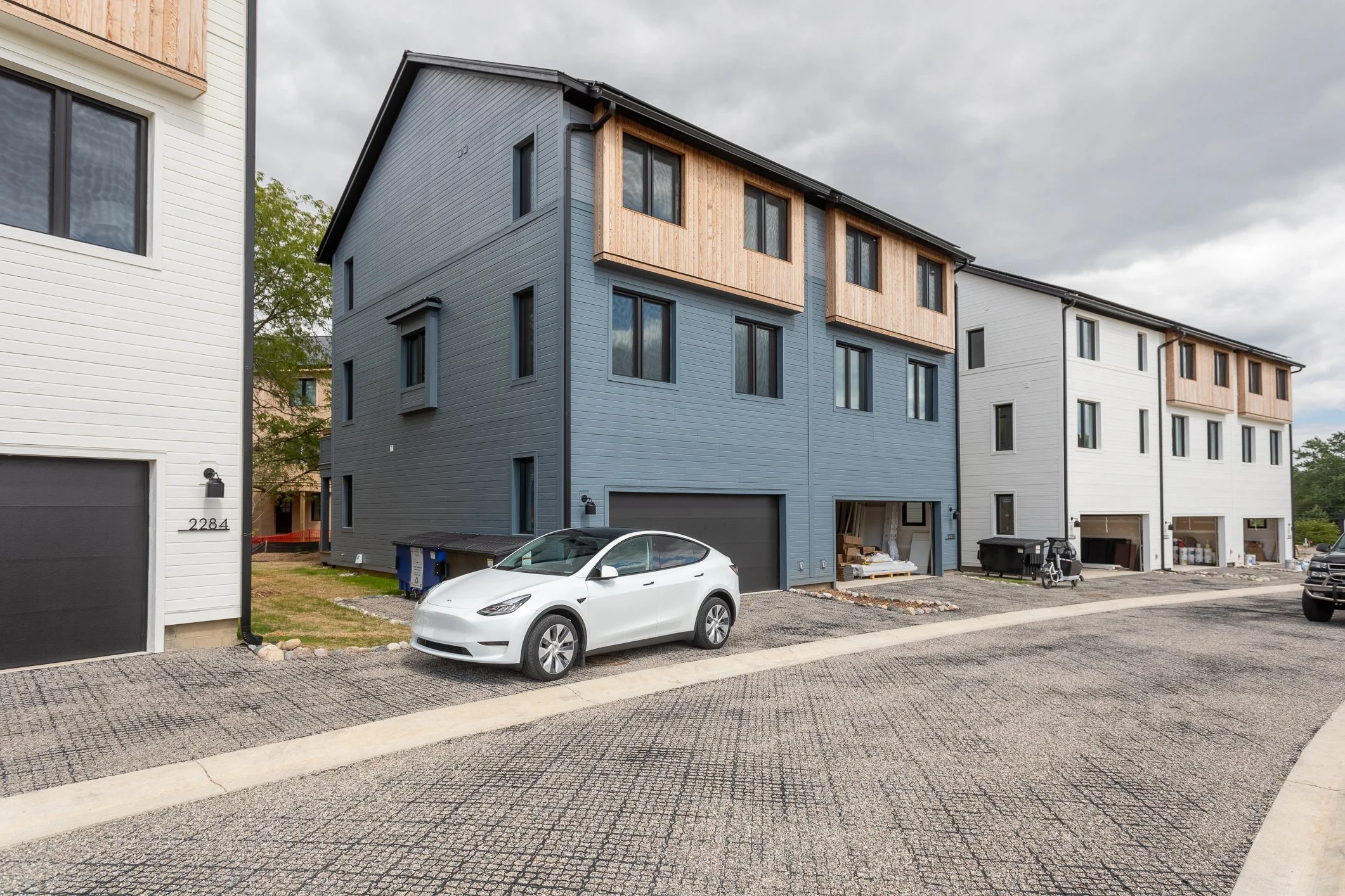 Newly constructed multi-story residential buildings with garage spaces on the ground floor, parked cars, construction materials, and equipment outside under an overcast sky.
