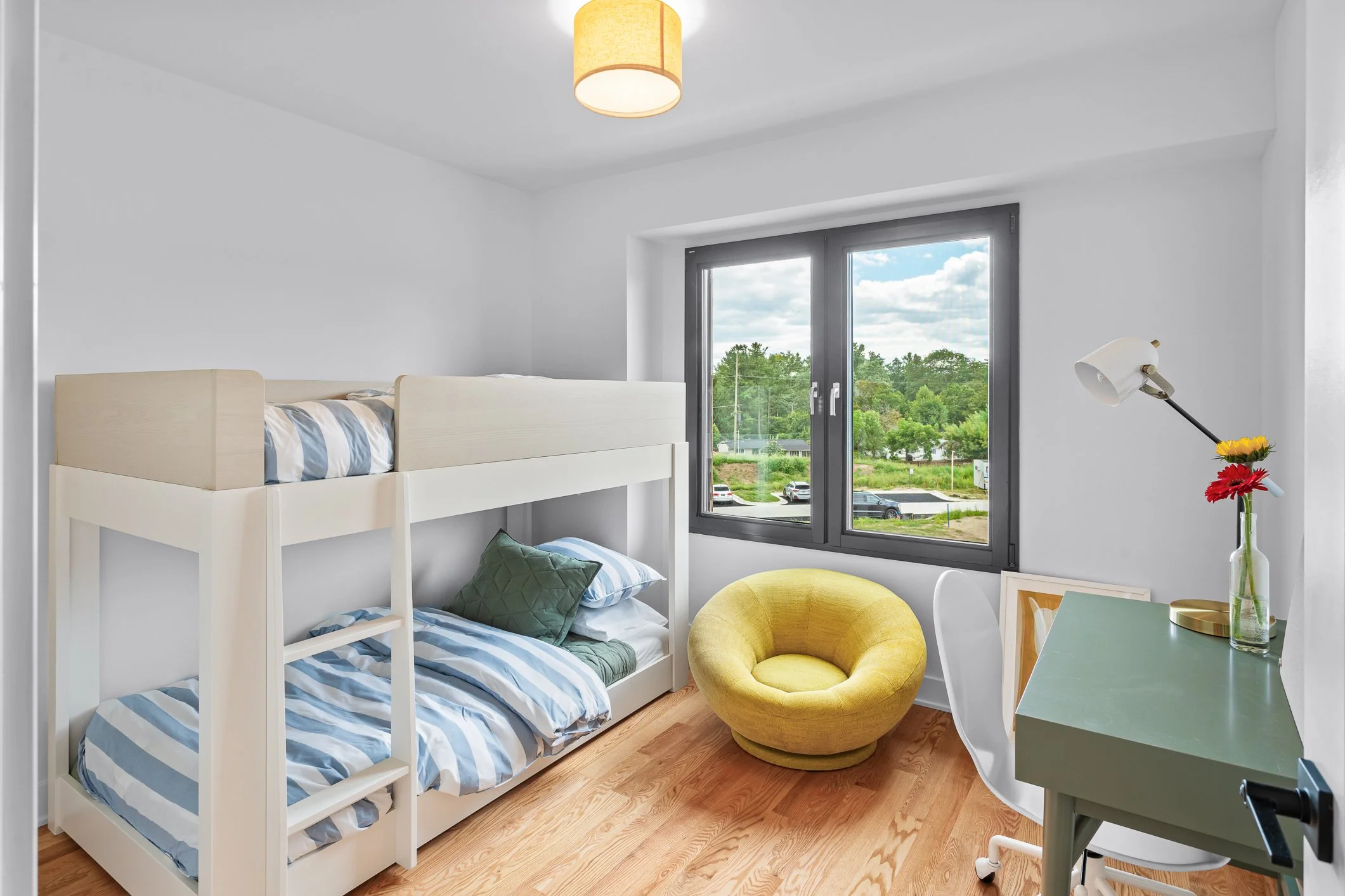 A bright bedroom with white walls and a bunk bed with blue and white striped bedding, a yellow round armchair, a desk with a white chair, and a window showing a view of trees and a parking lot.