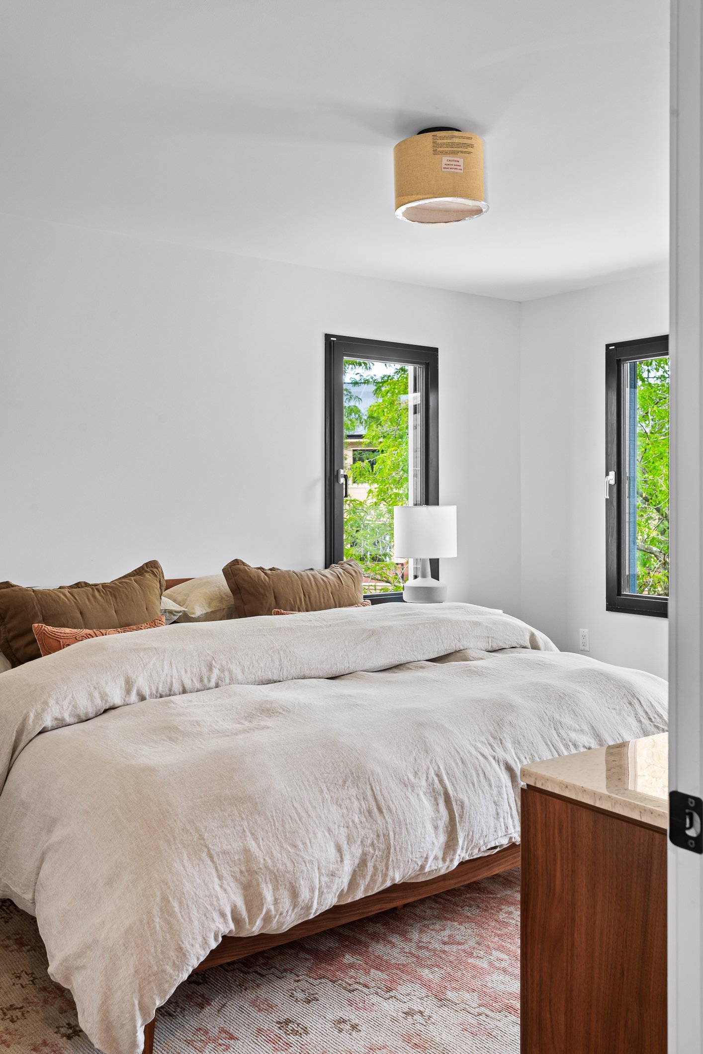 A bedroom with two windows showing green trees outside, a bed with beige and brown pillows, a white bedspread, a white lamp on a nightstand, and a ceiling light fixture.